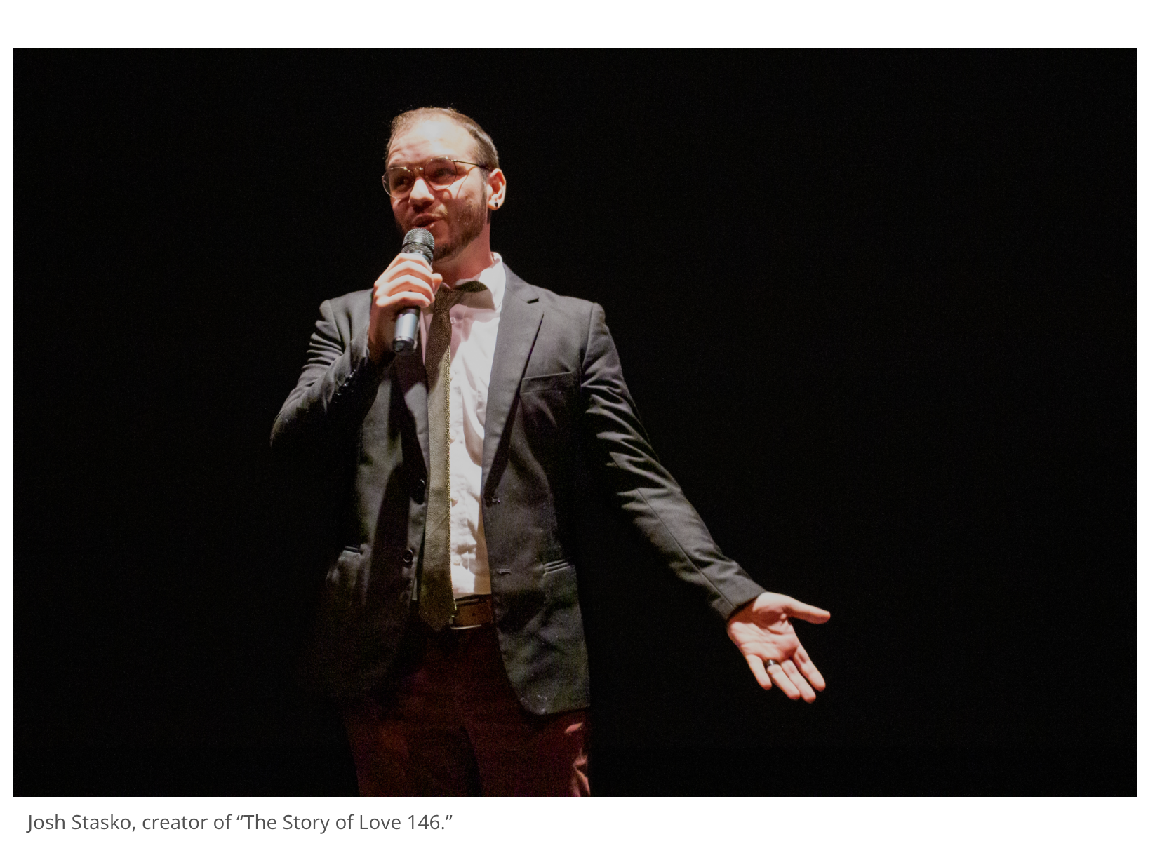 A man in a gray suit, white shirt, and glasses holding a microphone and gesturing with his left hand on a dark stage.