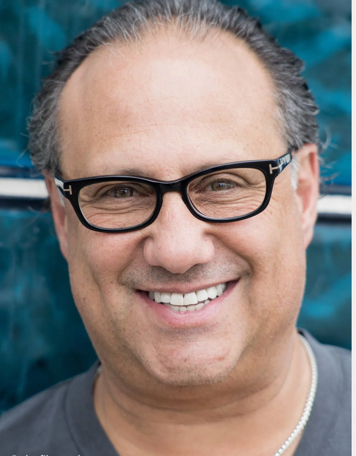 Close-up of a smiling man wearing black glasses, with gray hair, outdoors with a blue background.