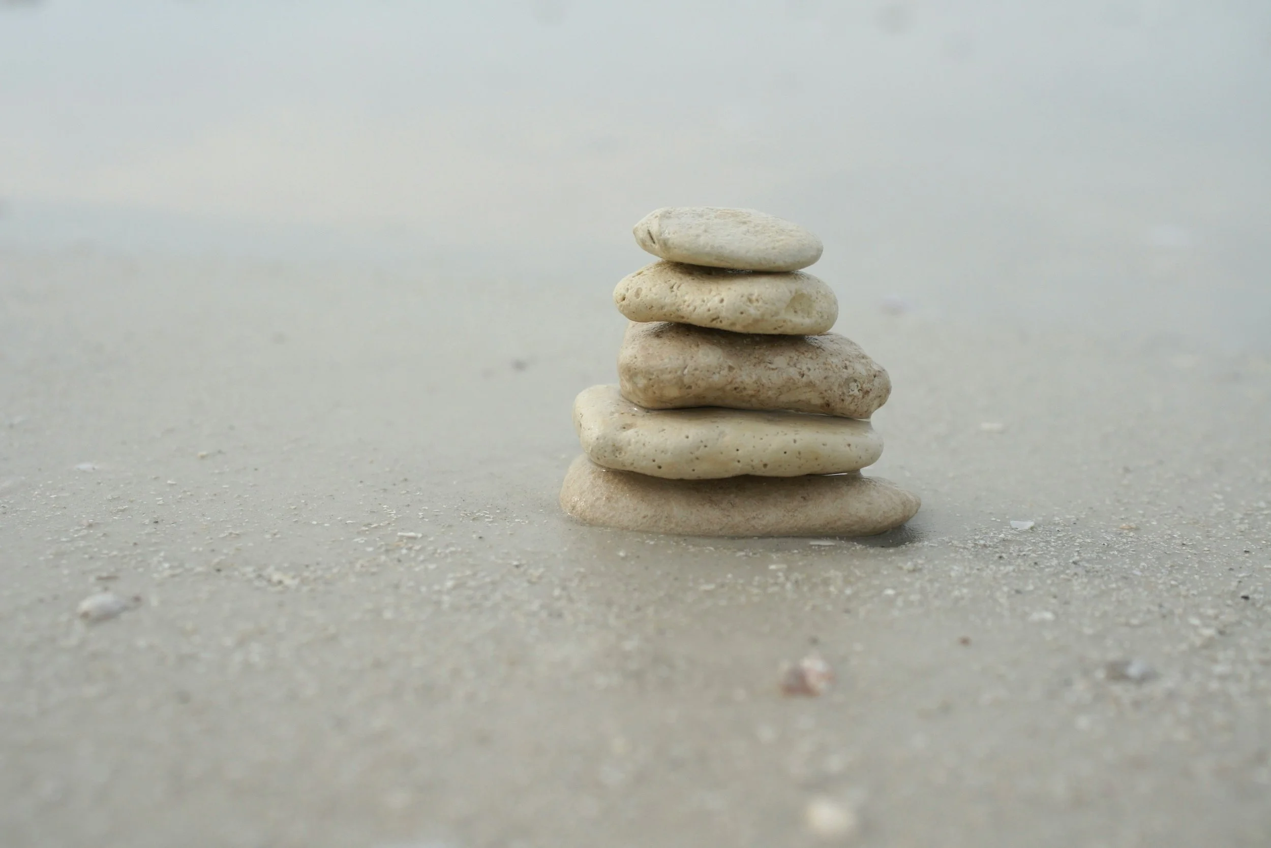 A small stack of five smooth, light-colored stones balanced on a sandy surface with a gray, cloudy sky in the background.