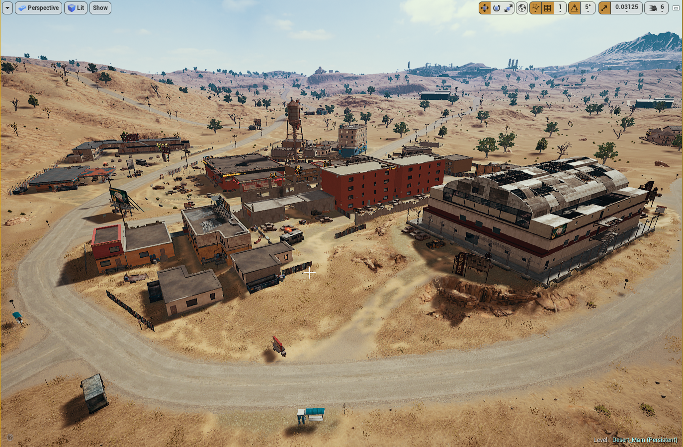 Desert landscape with a small, semi-urban settlement consisting of various buildings including a large dark red building, a warehouse, and smaller houses. There's a dirt road surrounding the settlement with sparse trees and hills in the background.