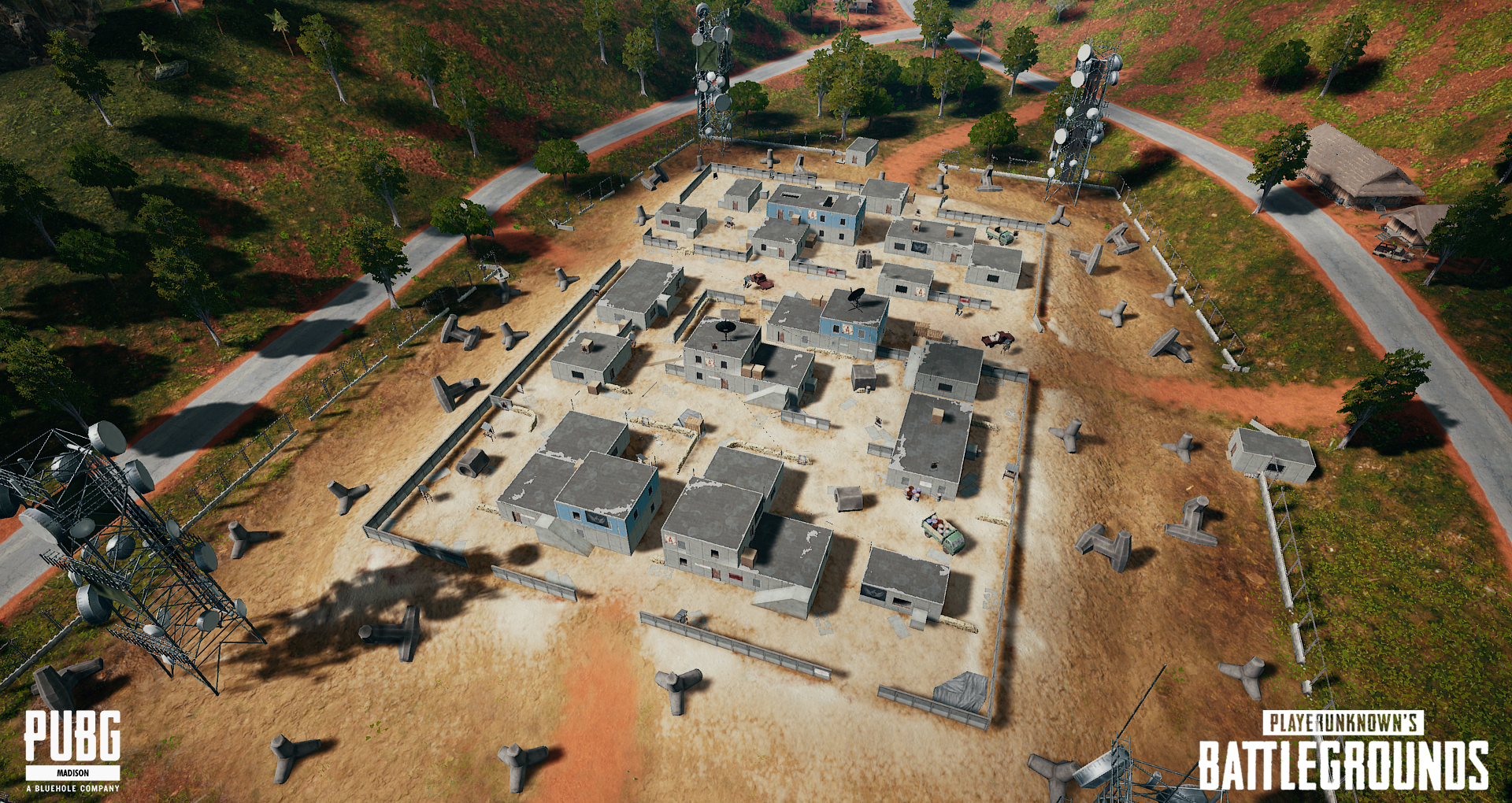 An aerial view of a small compound with multiple buildings surrounded by a chain-link fence, with dirt roads, scattered trees, and communication towers on red soil.