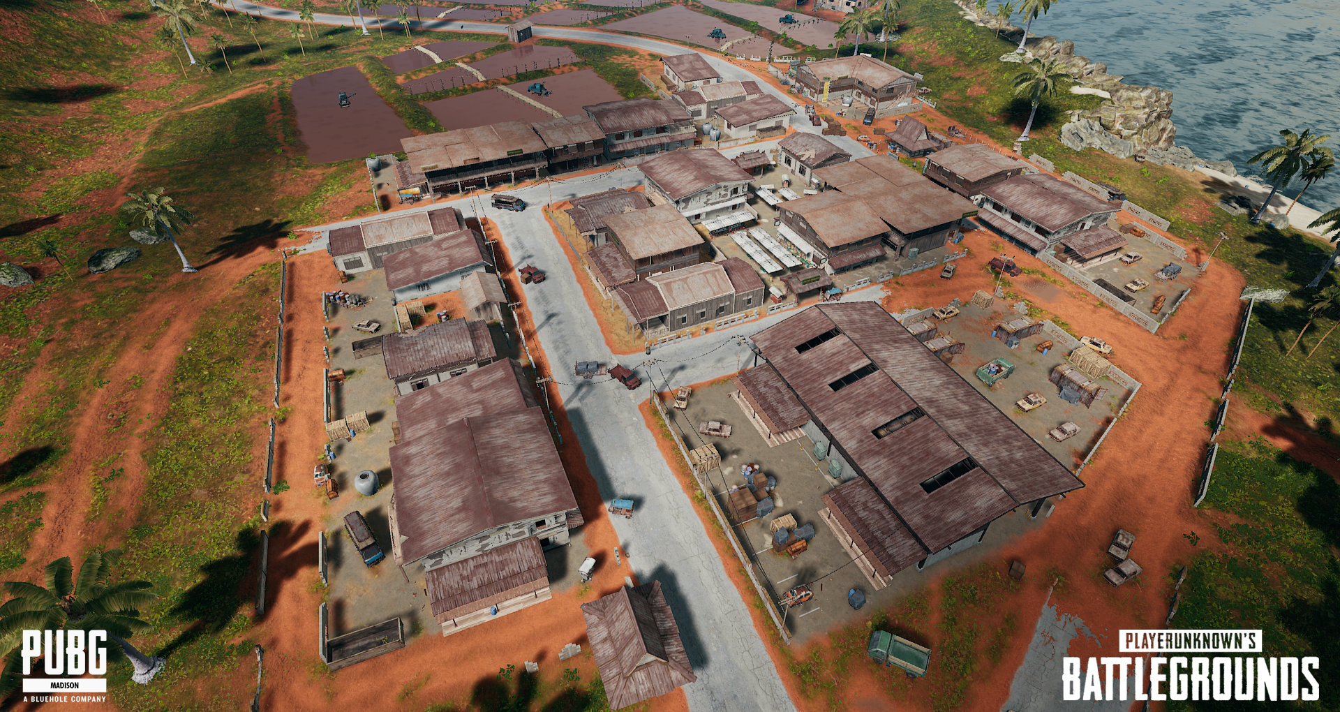 An aerial view of a rural compound with multiple buildings, roads, vehicles, and fenced areas, situated near water with rocks and palm trees.