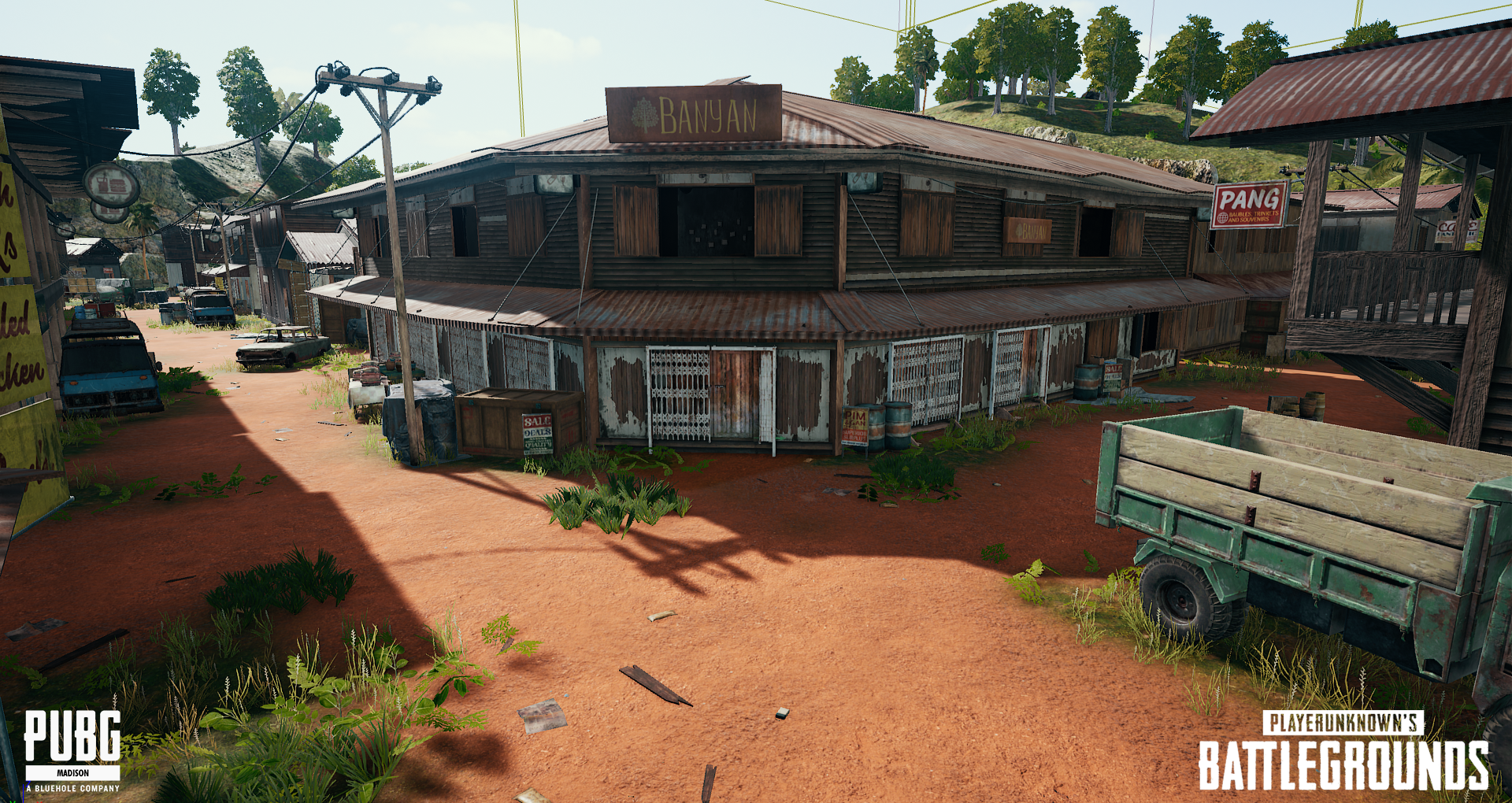 A rustic, weathered building with a sign that reads 'BANYAN,' surrounded by cars, barrels, and plants, in a rural game environment from PUBG Battlegrounds.