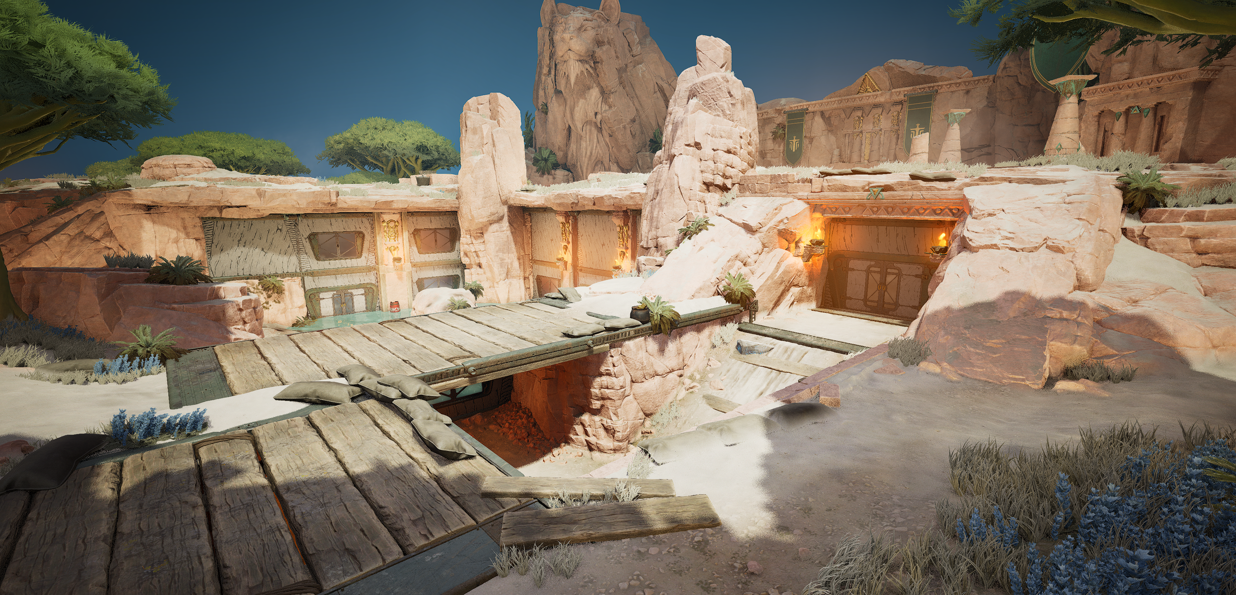 An ancient desert fortress carved into rocky cliffs, with wooden bridges connecting different levels and illuminated by torches, surrounded by sparse desert vegetation and large trees against a clear blue sky.