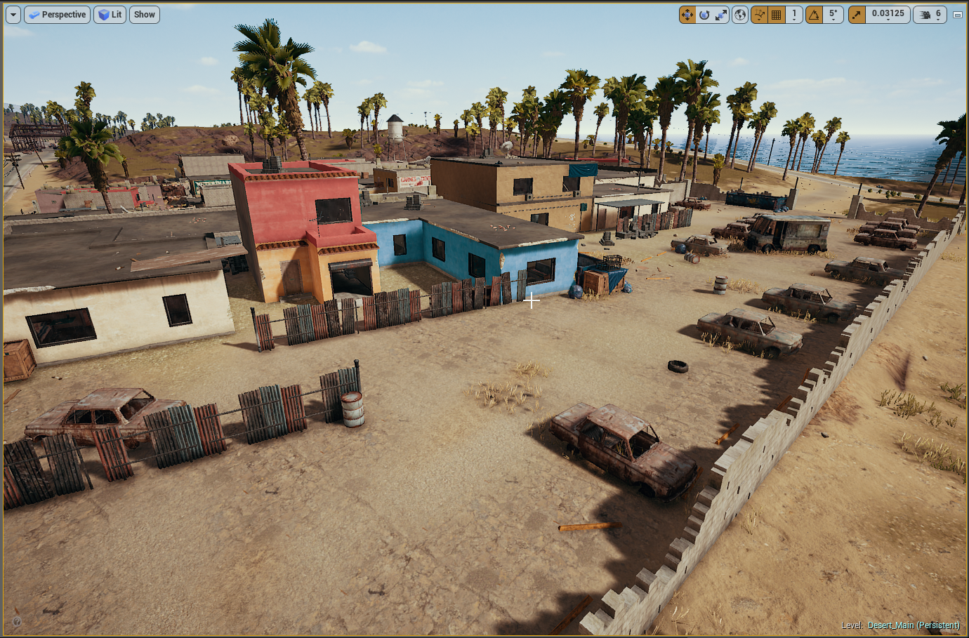 A dusty, semi-abandoned desert lot enclosed by a dirt wall, with old, rusty cars parked and scattered around. There are colorful small houses and huts in the background, some painted in bright pastel colors like blue, pink, and beige. Palm trees line