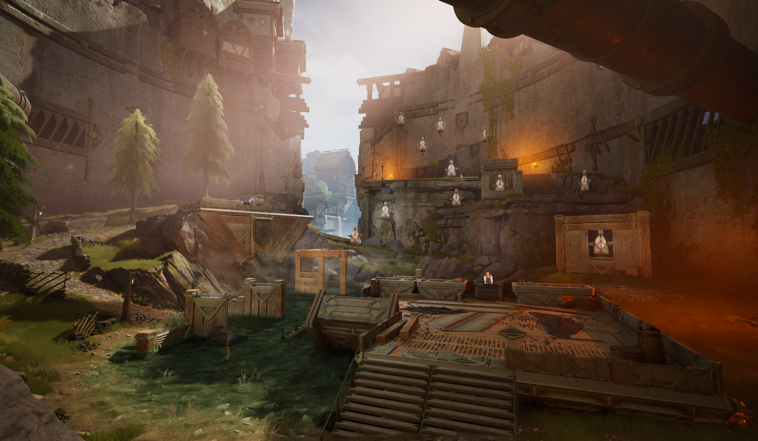 A fantasy video game scene showing a large, ancient fortress with wooden and stone structures set in a rocky landscape. There are trees on the left, a small river, and crates or containers scattered around. The scene is illuminated by warm lighting, 