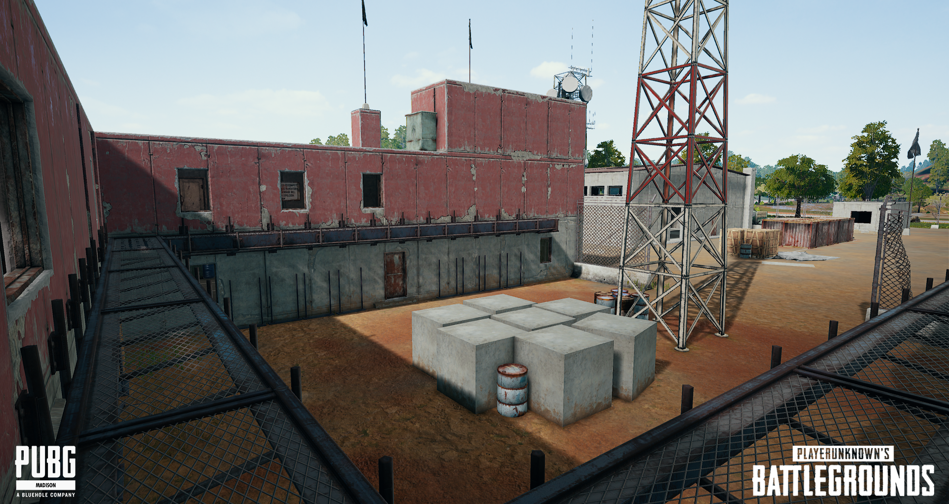 From the elevated perspective, an outdoor training or industrial area with a red weathered building, a dirt ground, large concrete blocks, a barrel, and hanging tires, enclosed by a chain-link fence and a tall steel tower, with trees and clear sky in