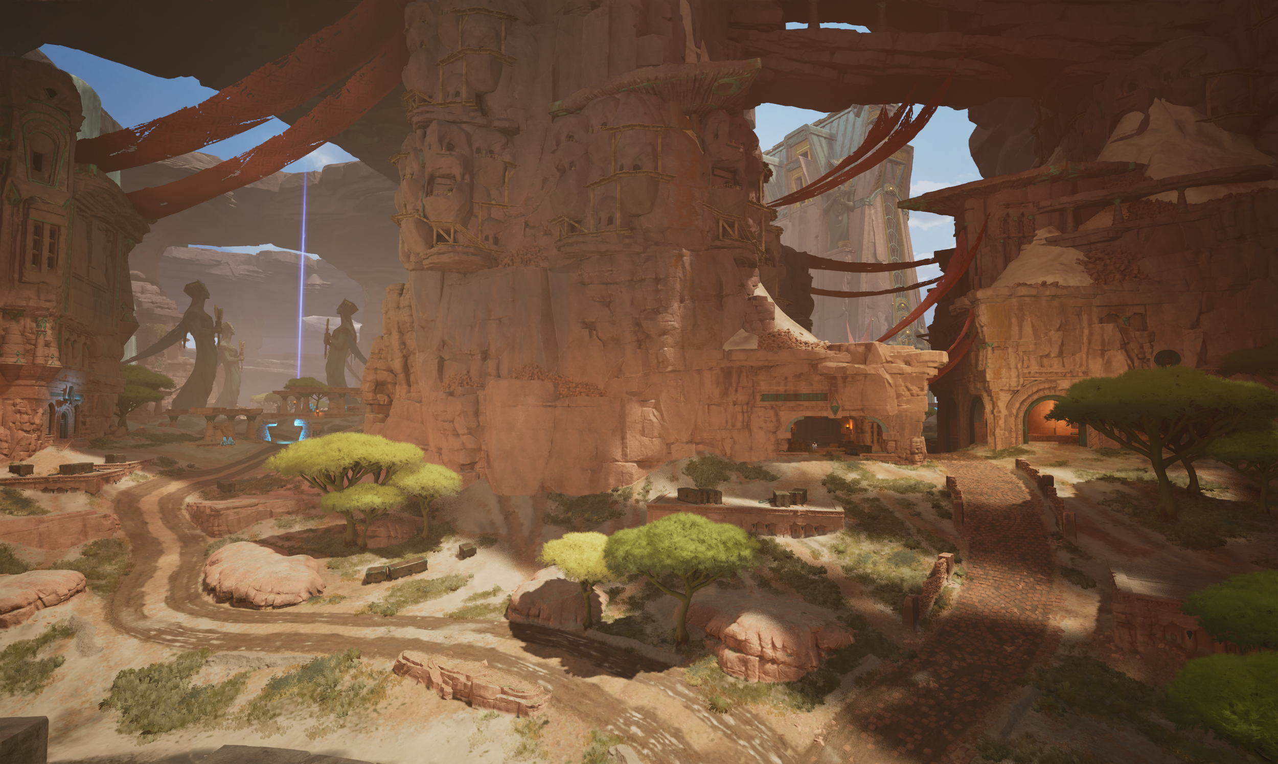 A desert landscape with large reddish rock formations, winding dirt paths, and scattered green trees. There are structures built into the rock formations and a tall stone tower visible in the background.