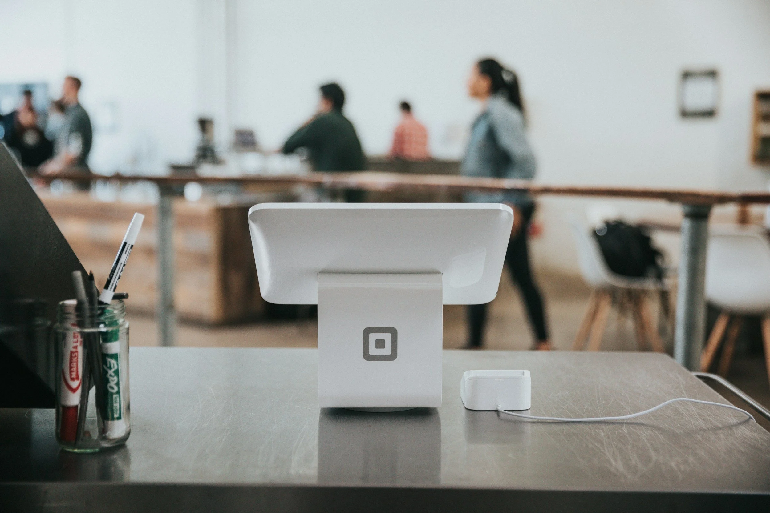 A modern retail checkout counter with a white cash register, a small white card reader connected by a wire, and a jar of markers on a gray counter, with blurred customers and a wooden railing in the background.