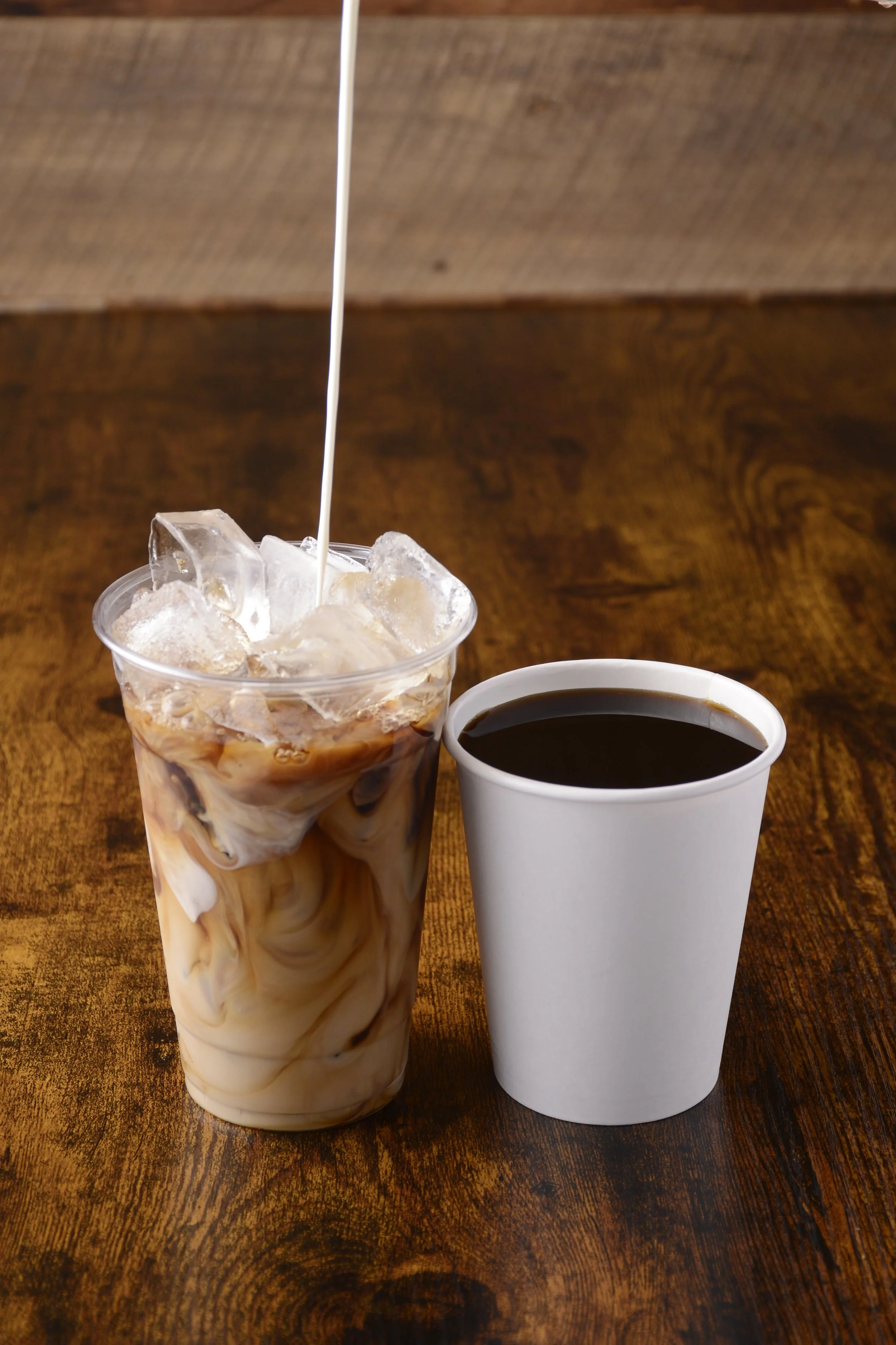 A clear cup of iced coffee with swirls of milk and ice beside a paper cup of black coffee