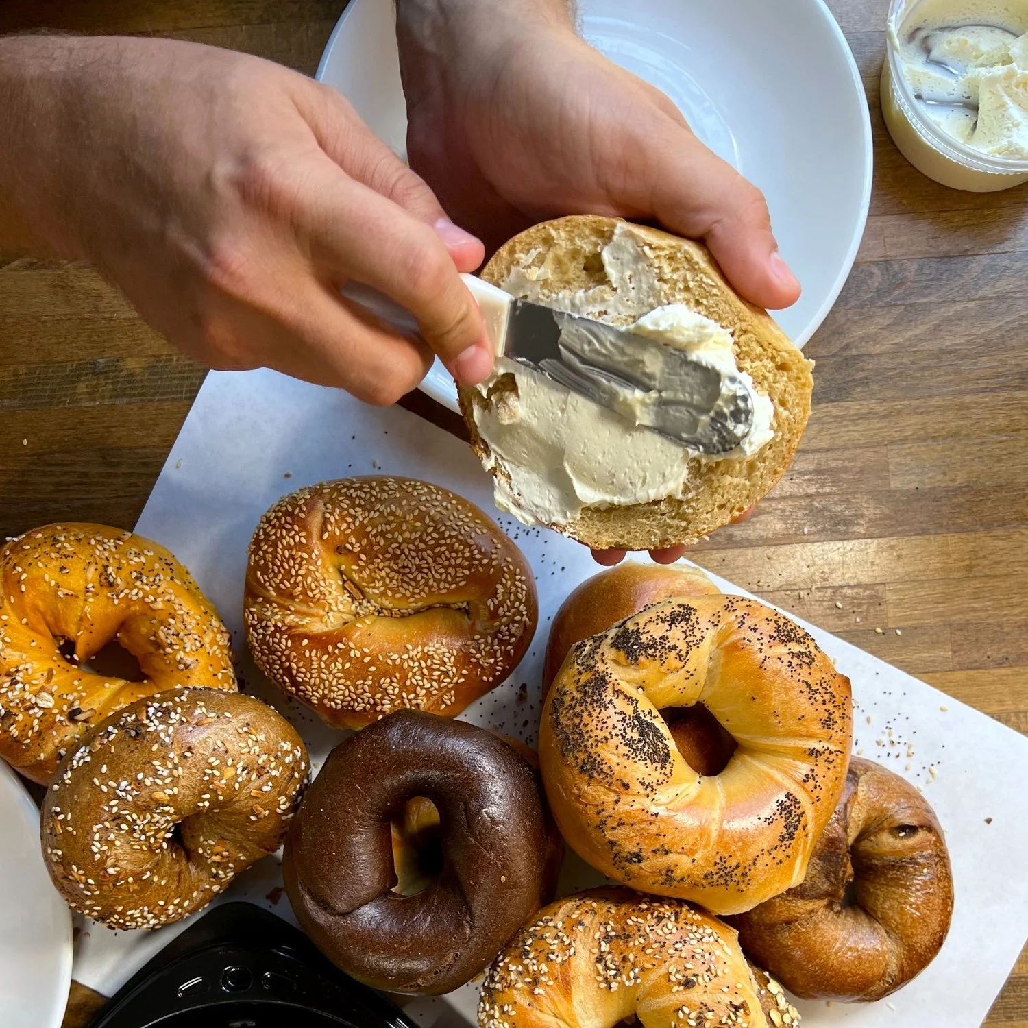 Person spreading cream cheese on a bagel with various bagels on a tray below.