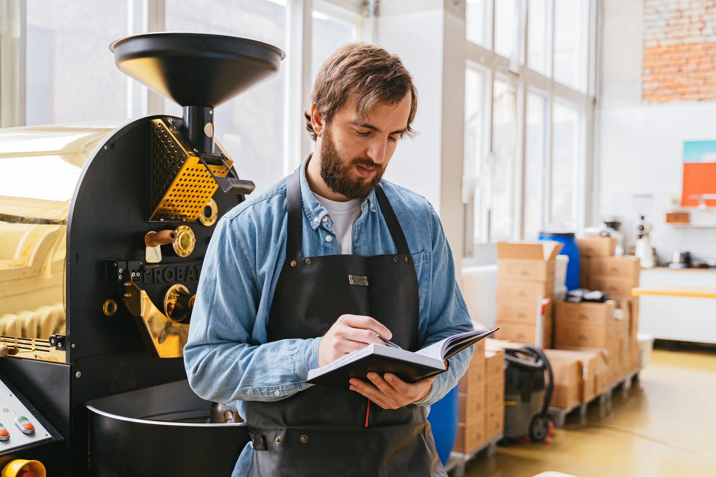 A man with a beard wearing a denim shirt and black apron, standing in a workspace, looking at a notebook and writing notes.