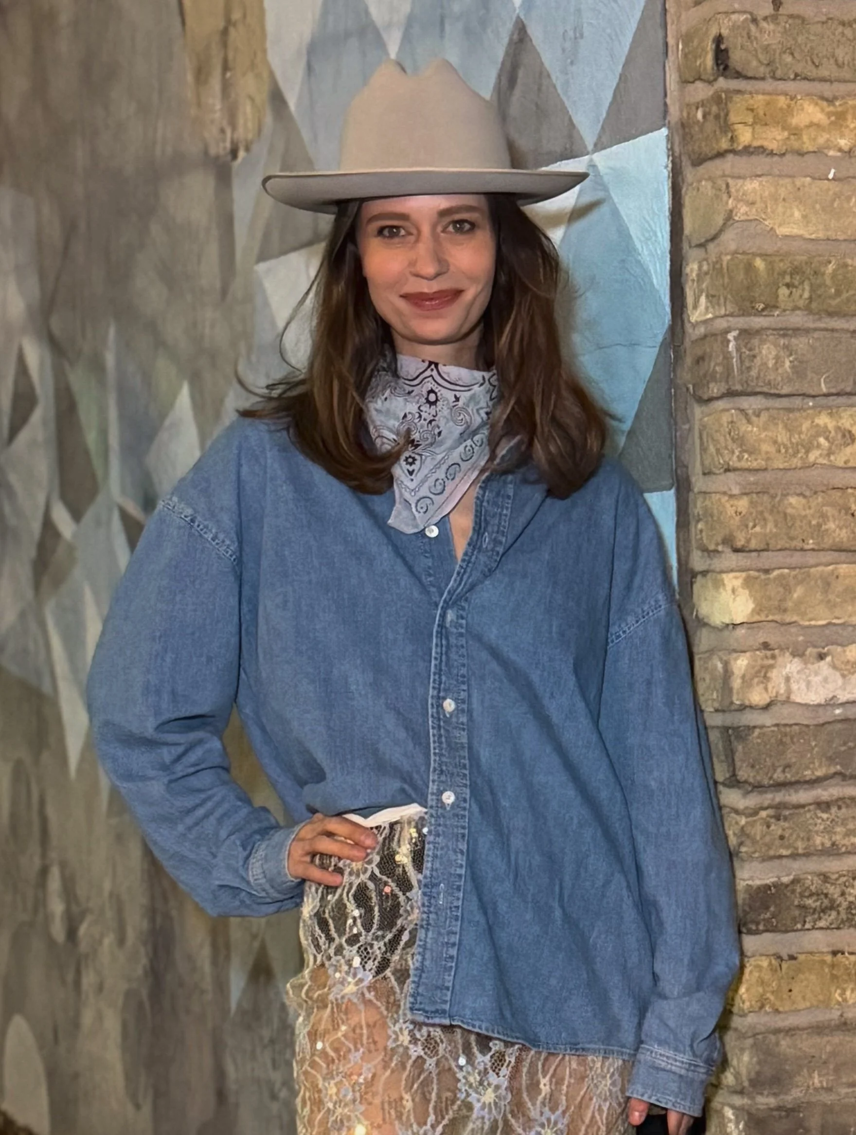 A woman with long brown hair wearing a beige cowboy hat, denim shirt, white bandana with black patterns around her neck, holding a small purse with snakeskin pattern, standing between a stone wall and brick wall.