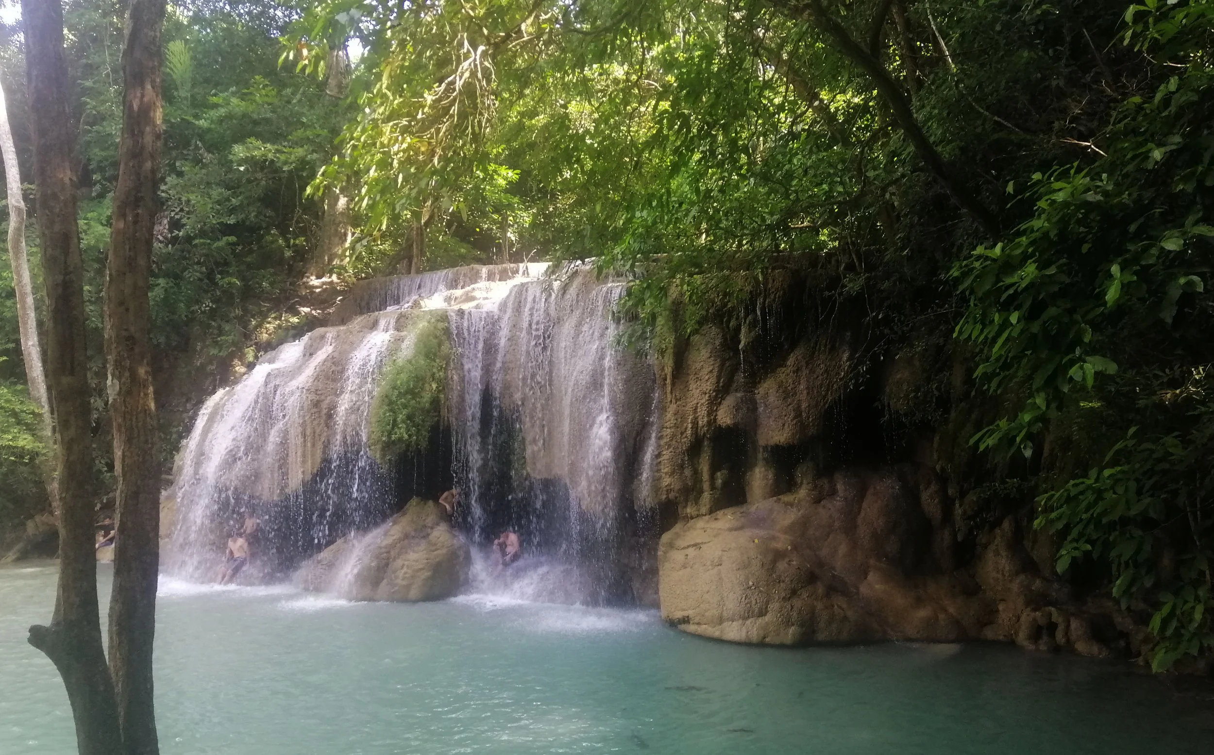 The image shows the famous Erawan Waterfall located in Erawan National Park in the Kanchanaburi Province of western Thailand. The turquoise color of the water is caused by the high mineral content, specifically calcium carbonate, depositing on the ro