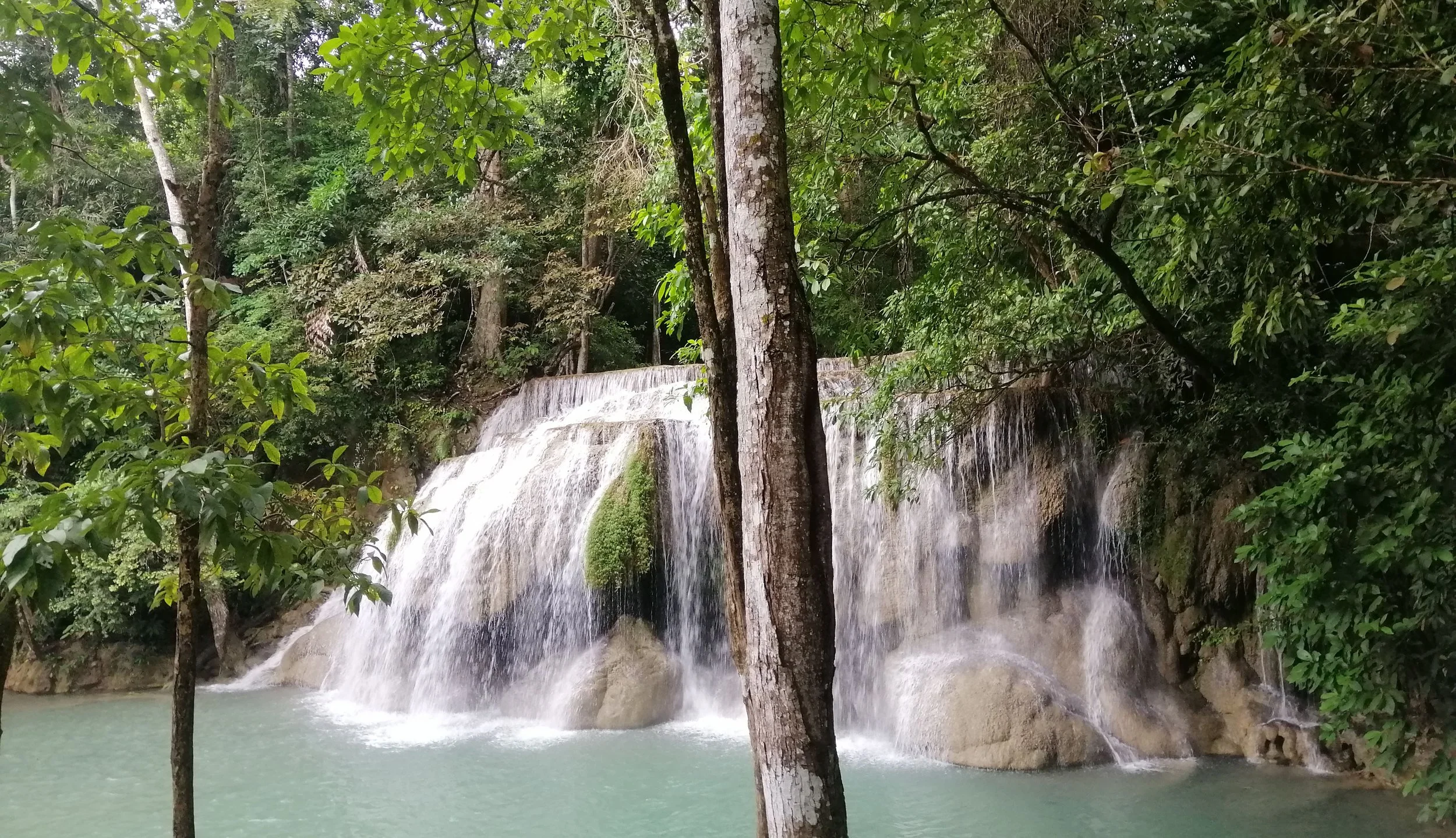 The image shows the famous Erawan Waterfall located in Erawan National Park in the Kanchanaburi Province of western Thailand.  The falls are composed of massive limestone and dolomitic limestone, which often feature chert nodules and marine fossils. 