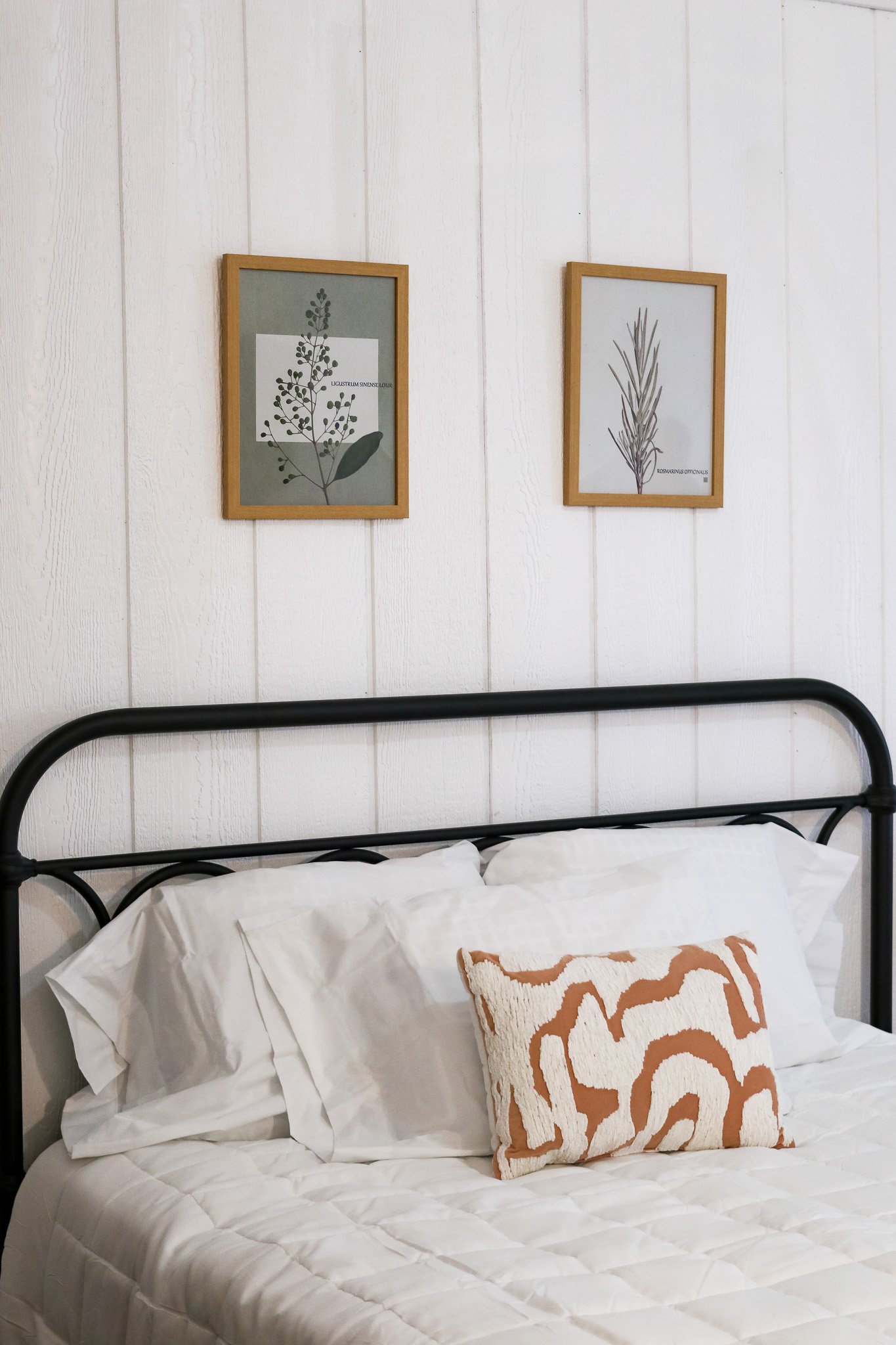 Minimalist bedroom with a metal headboard, two framed plant illustrations on the wall, and a white bedding set with a patterned decorative pillow.