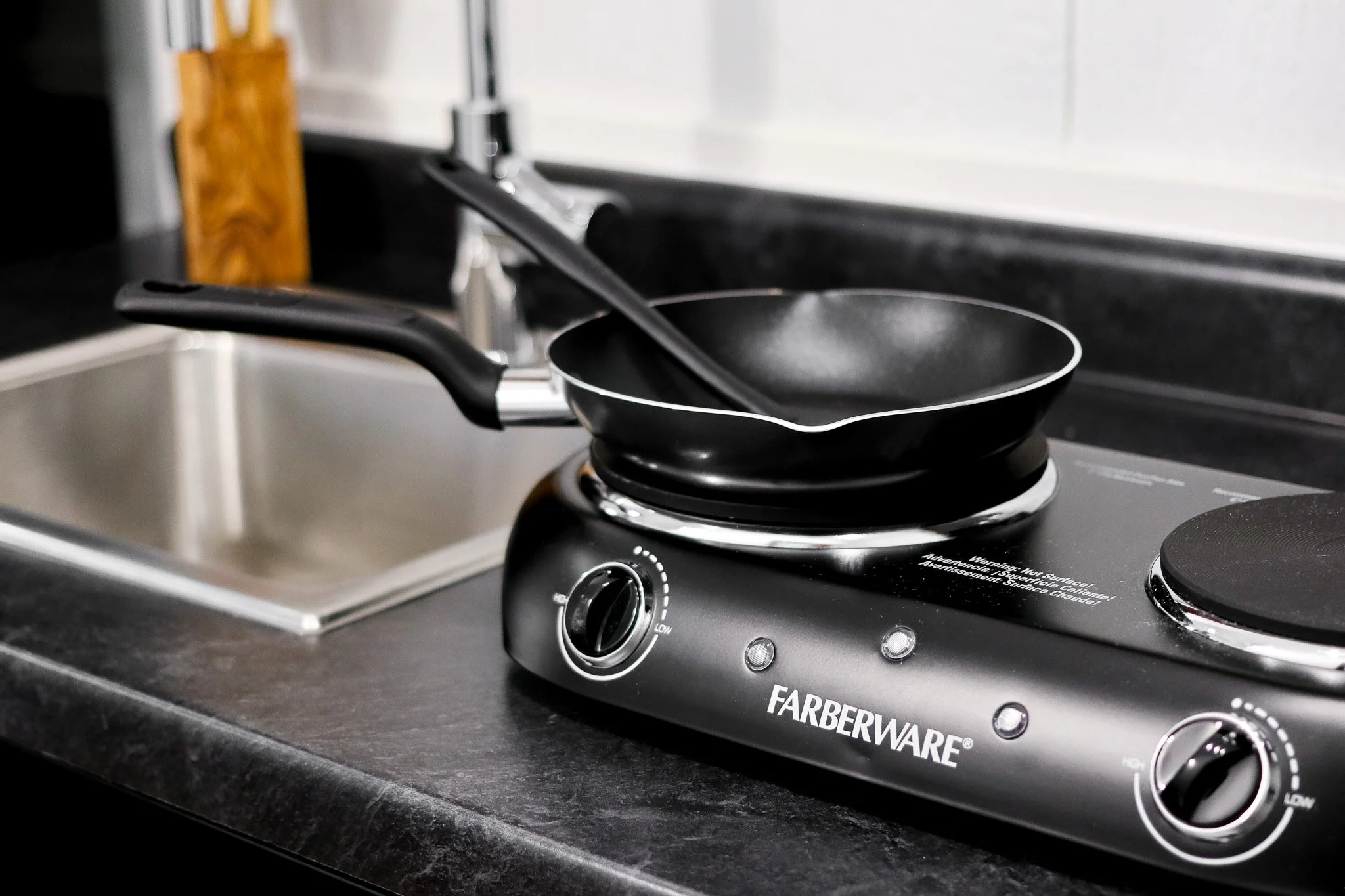 Electric countertop hot plate with a frying pan on it next to a kitchen sink.