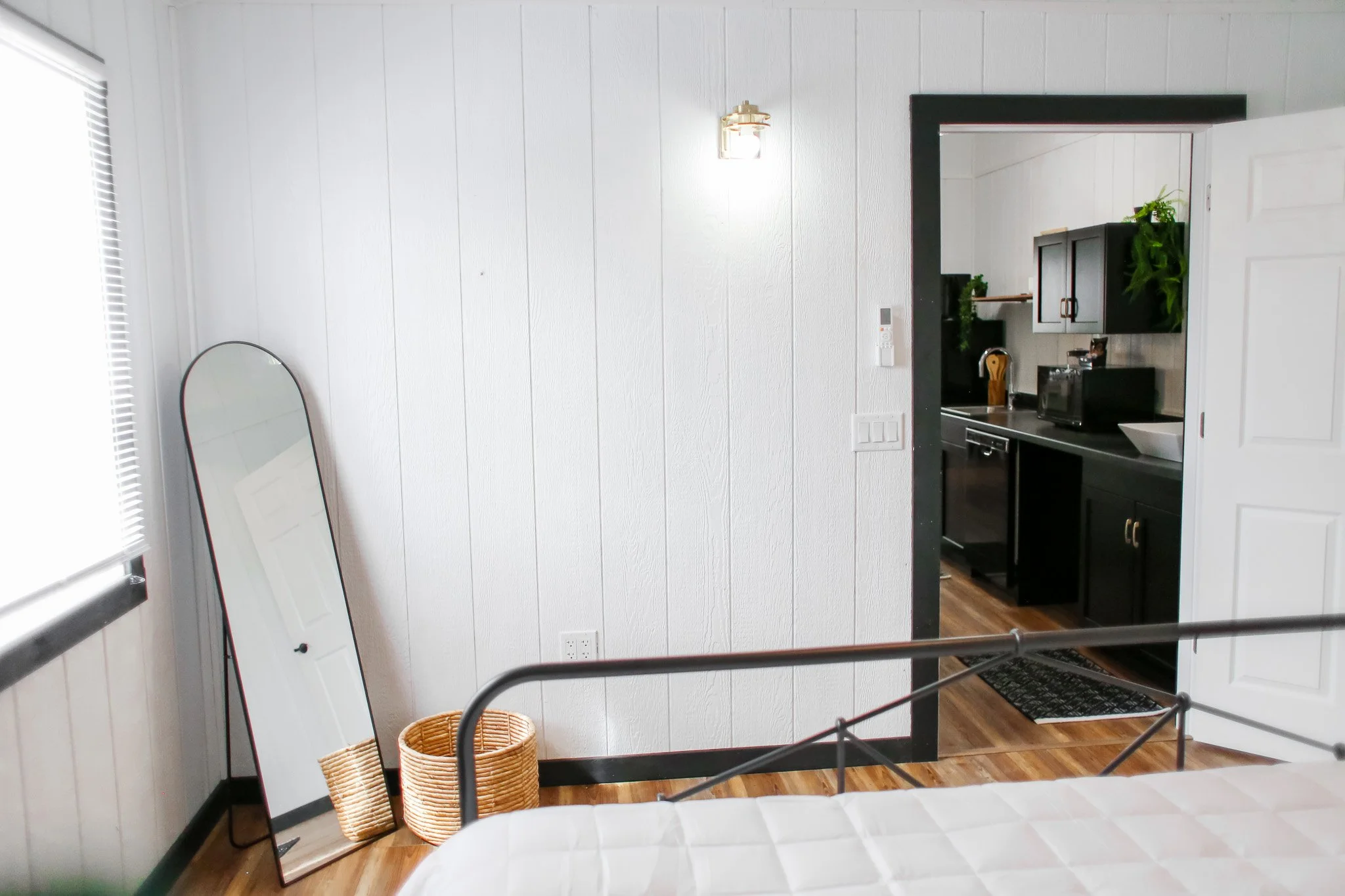 Minimalist bedroom with a standing mirror, wicker basket, and view into a kitchen.