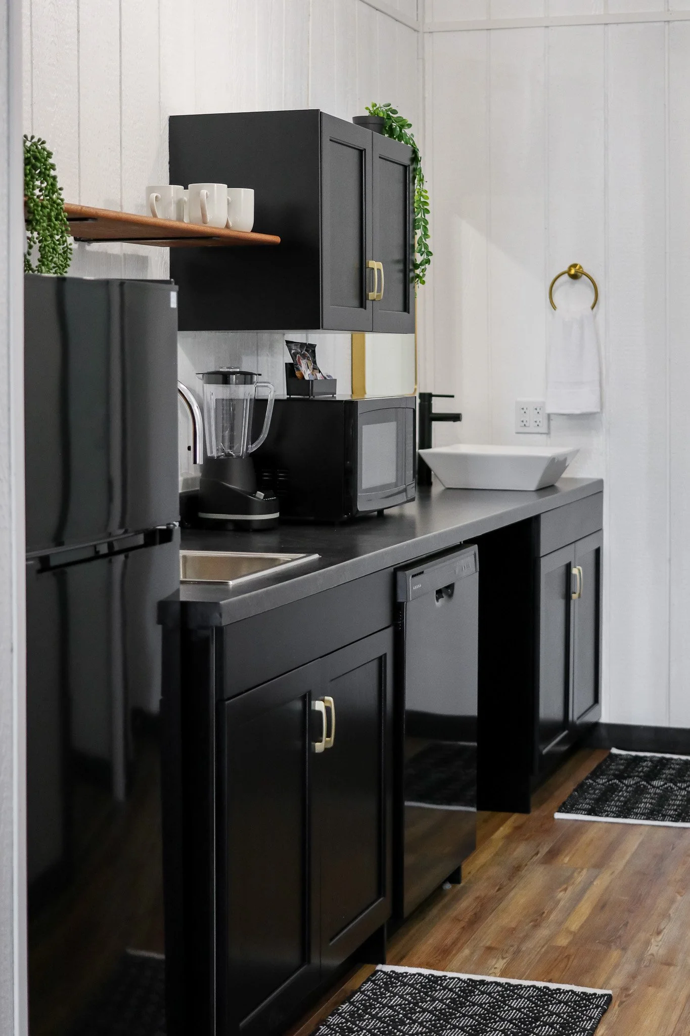 Modern kitchen with black cabinets, appliances, wooden shelves, plants, sink, and a towel holder on a white wall.