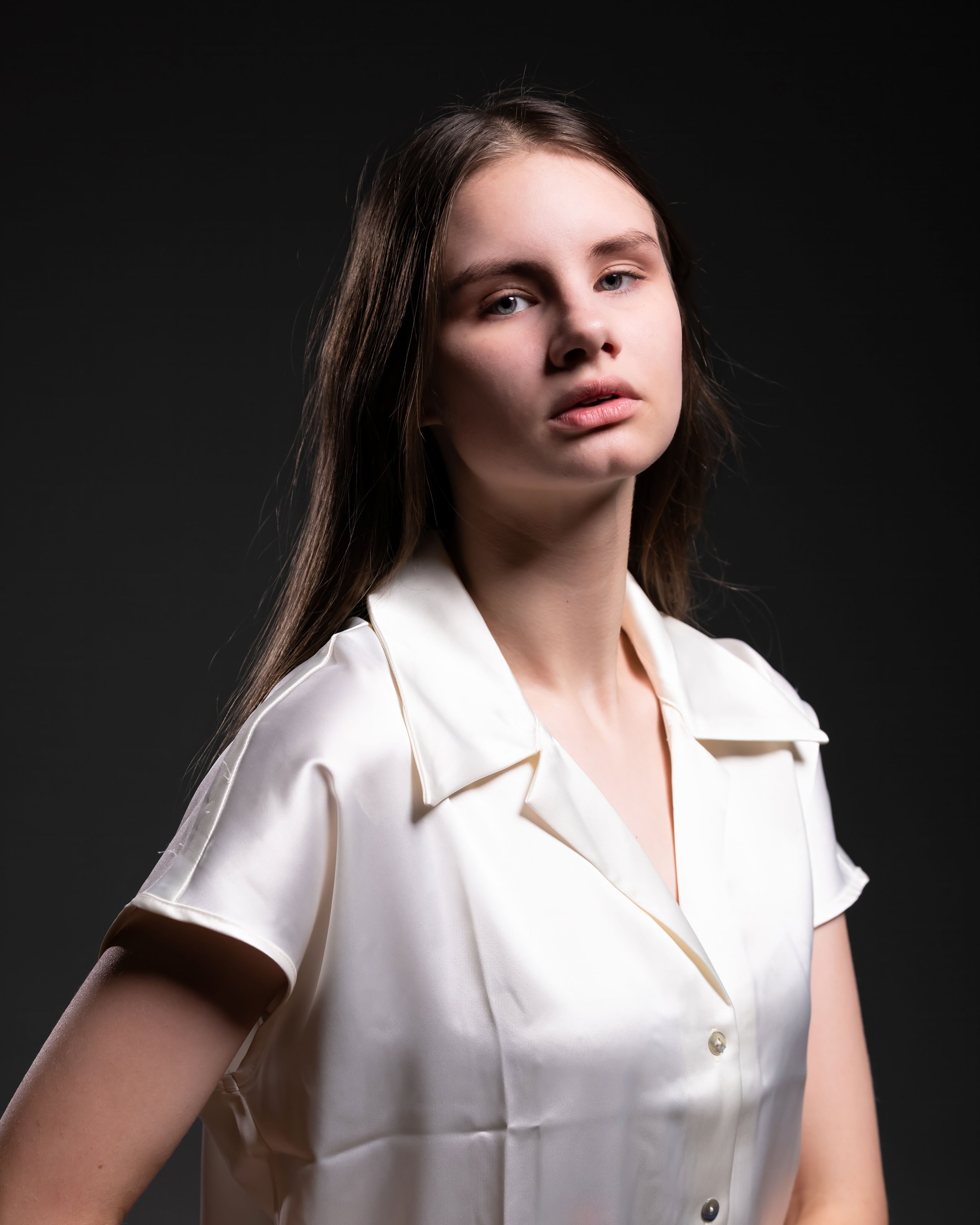 Portrait headshot of woman in a white short sleeved blouse. Lighting is dark and moody. The woman has a serious expression on her face. Fremont High School Dance Company