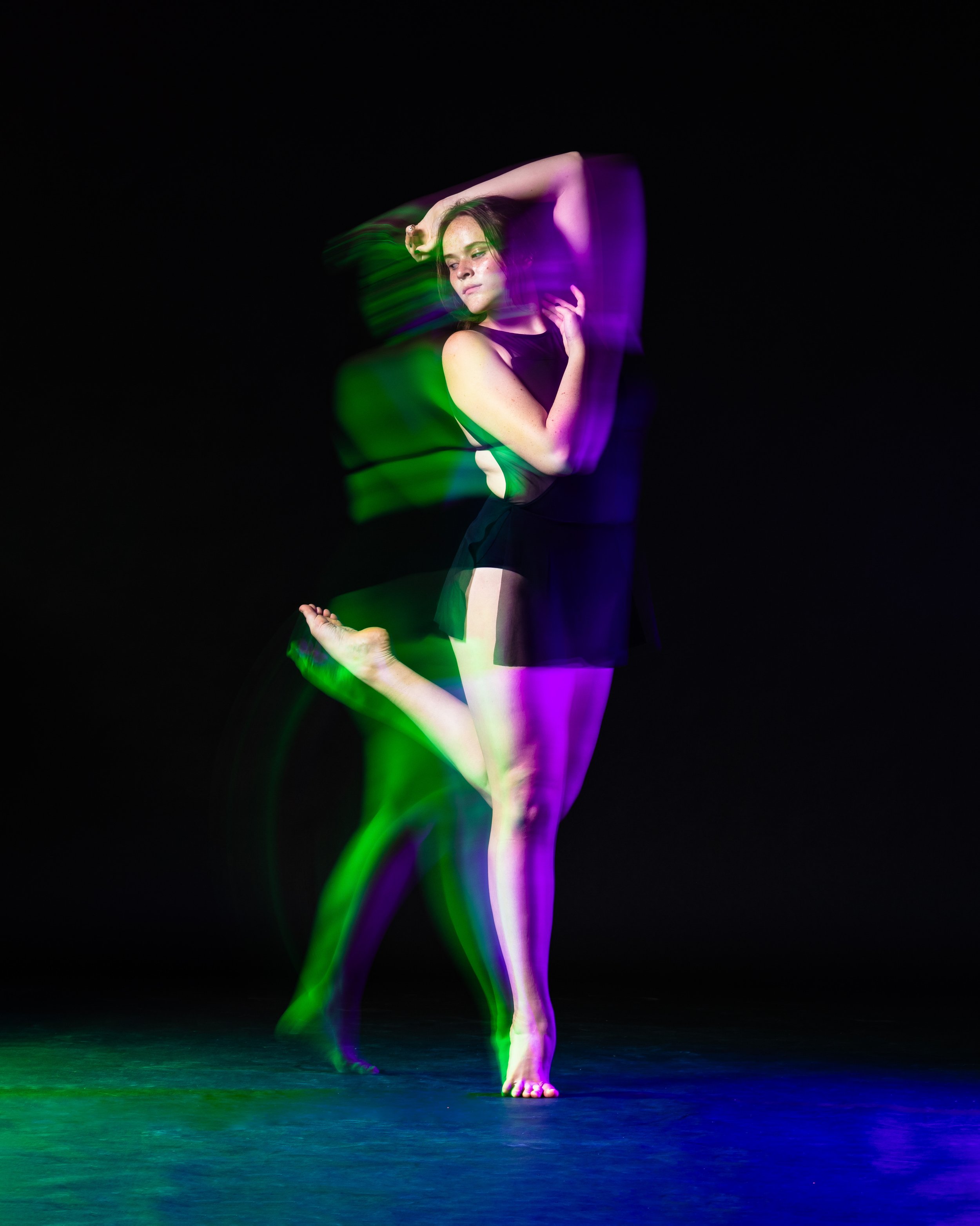 Dance photographer image of long exposure. Dancer has green and purple light trails. Dancer is framing her face with her hands while kicking her leg on releve. Dancer is looking at back foot. Davis High School Dance Company