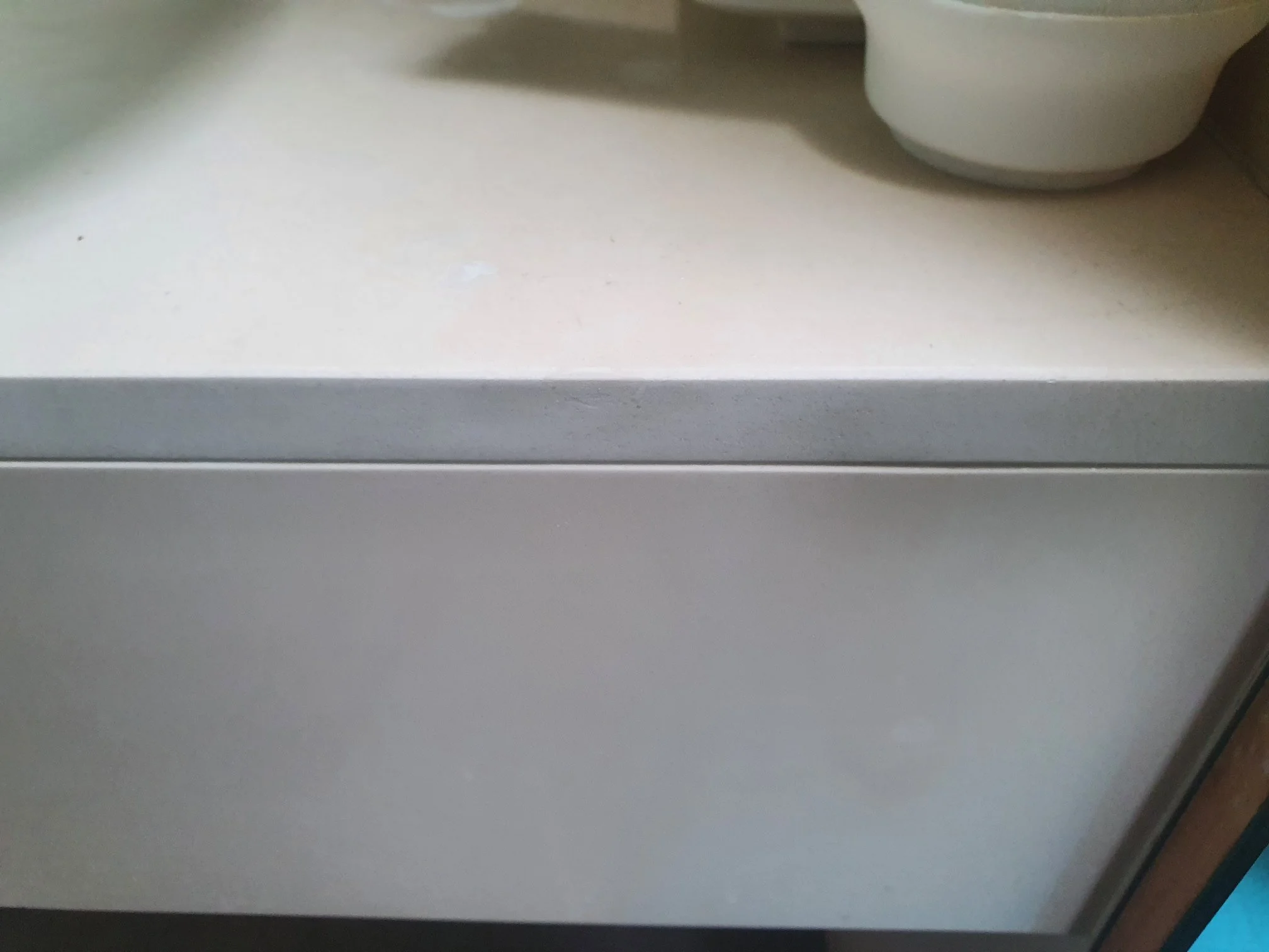 Close-up of a white kitchen cabinet or drawer with a round white cup or mug placed on top, against a light-colored countertop.