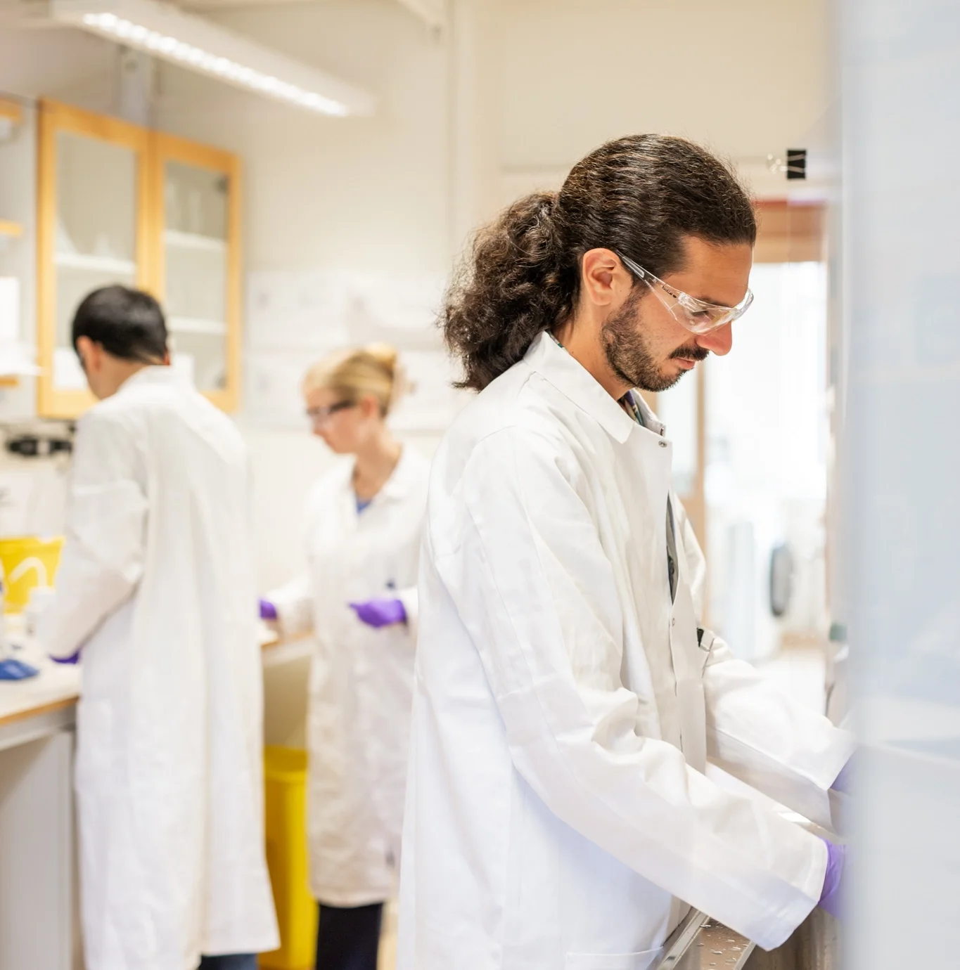 Researcher in a lab coat and safety glasses working at a fume hood, with two colleagues working at lab benches in the background.