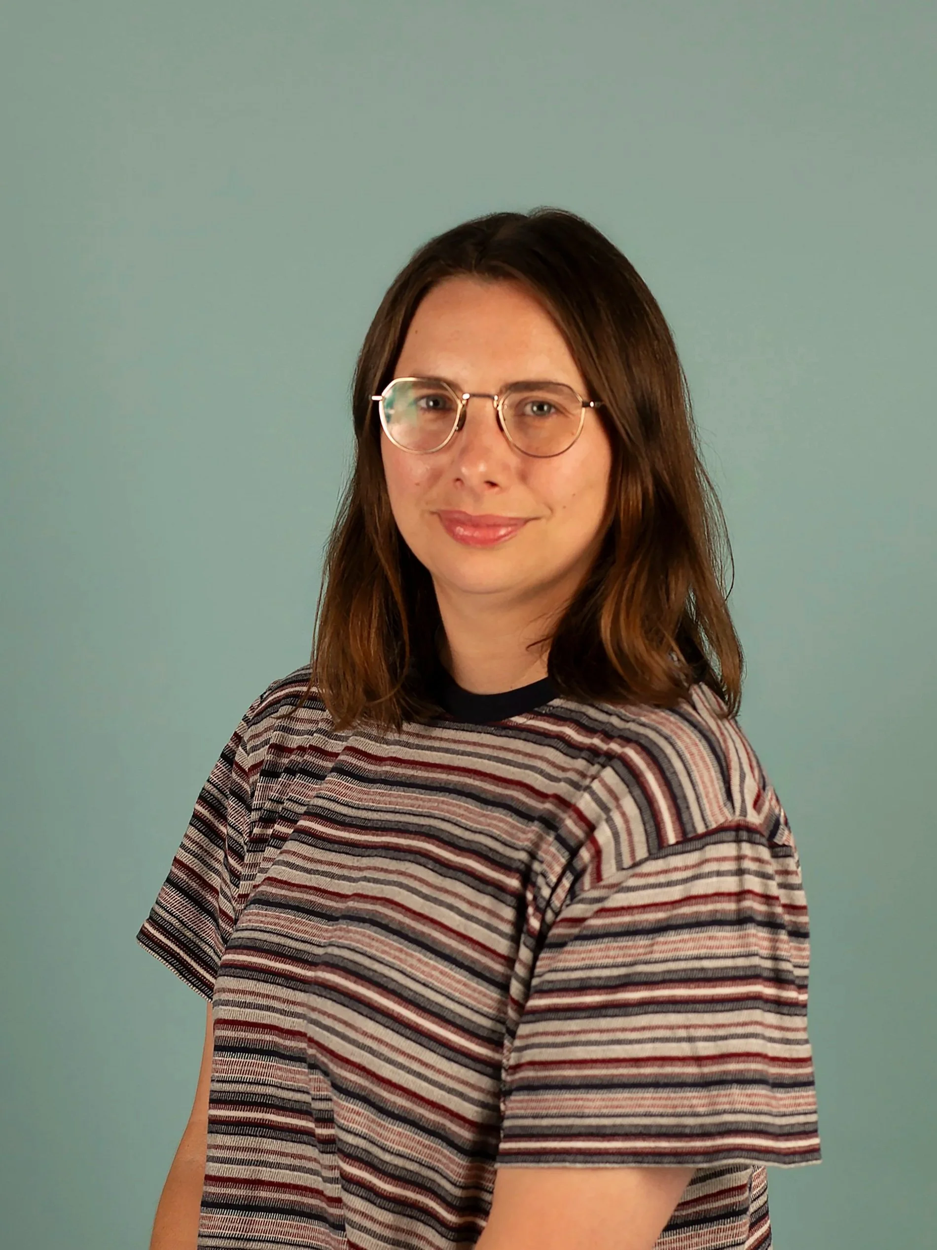 A woman with shoulder-length brown hair, wearing round glasses and a striped t-shirt, standing against a plain light teal background.