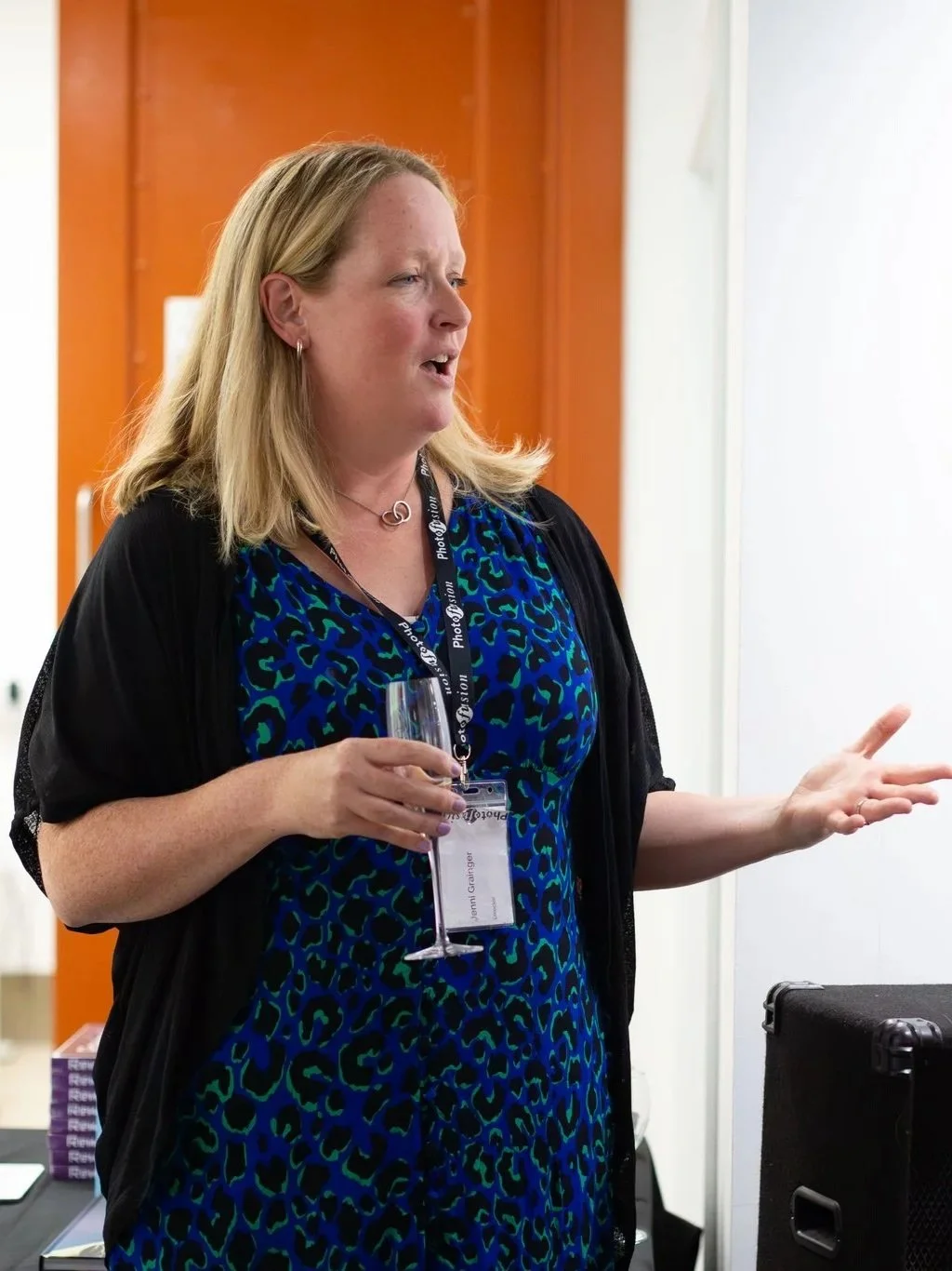 Woman speaking at an event, holding a wine glass, wearing a name badge, black cardigan, and a blue and green patterned dress.