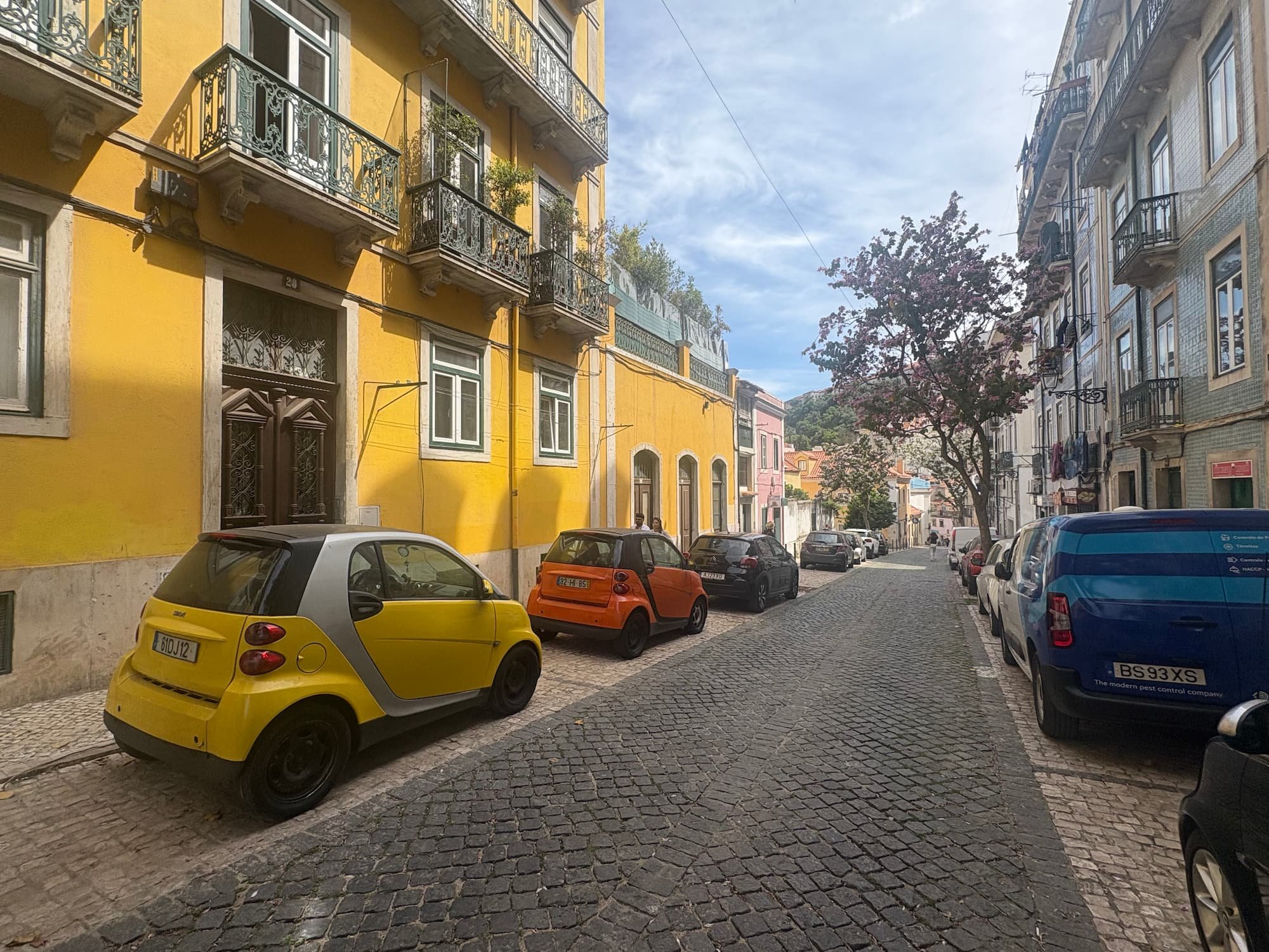 Colourful cobbled street in Lisbon's Alfama district, lined with traditional Portuguese buildings and parked cars, showcasing the neighbourhood's historic charm.