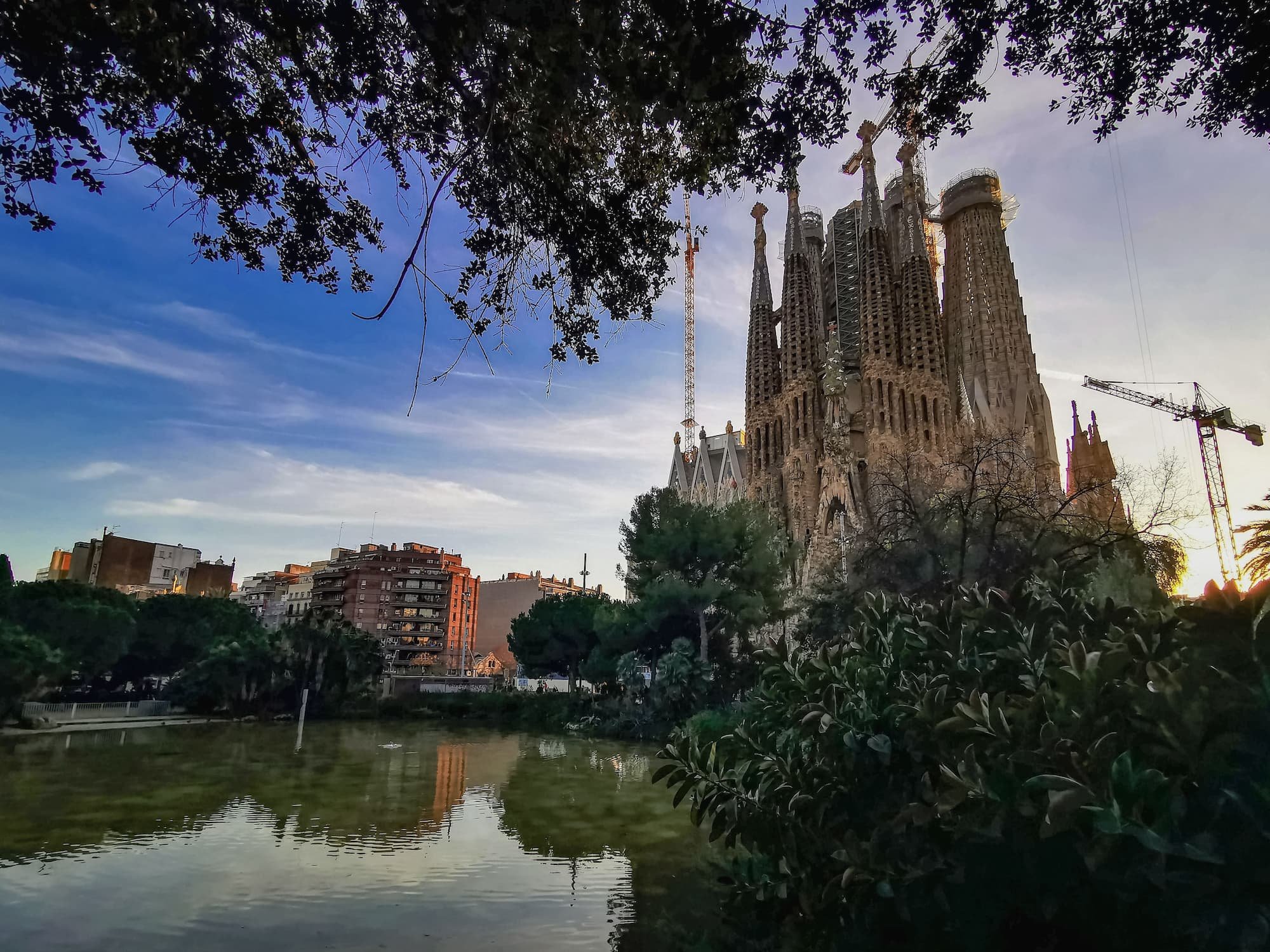 La Sagrada Família in Barcelona at sunset, viewed across a pond with reflections of Antoni Gaudí's iconic basilica surrounded by trees.