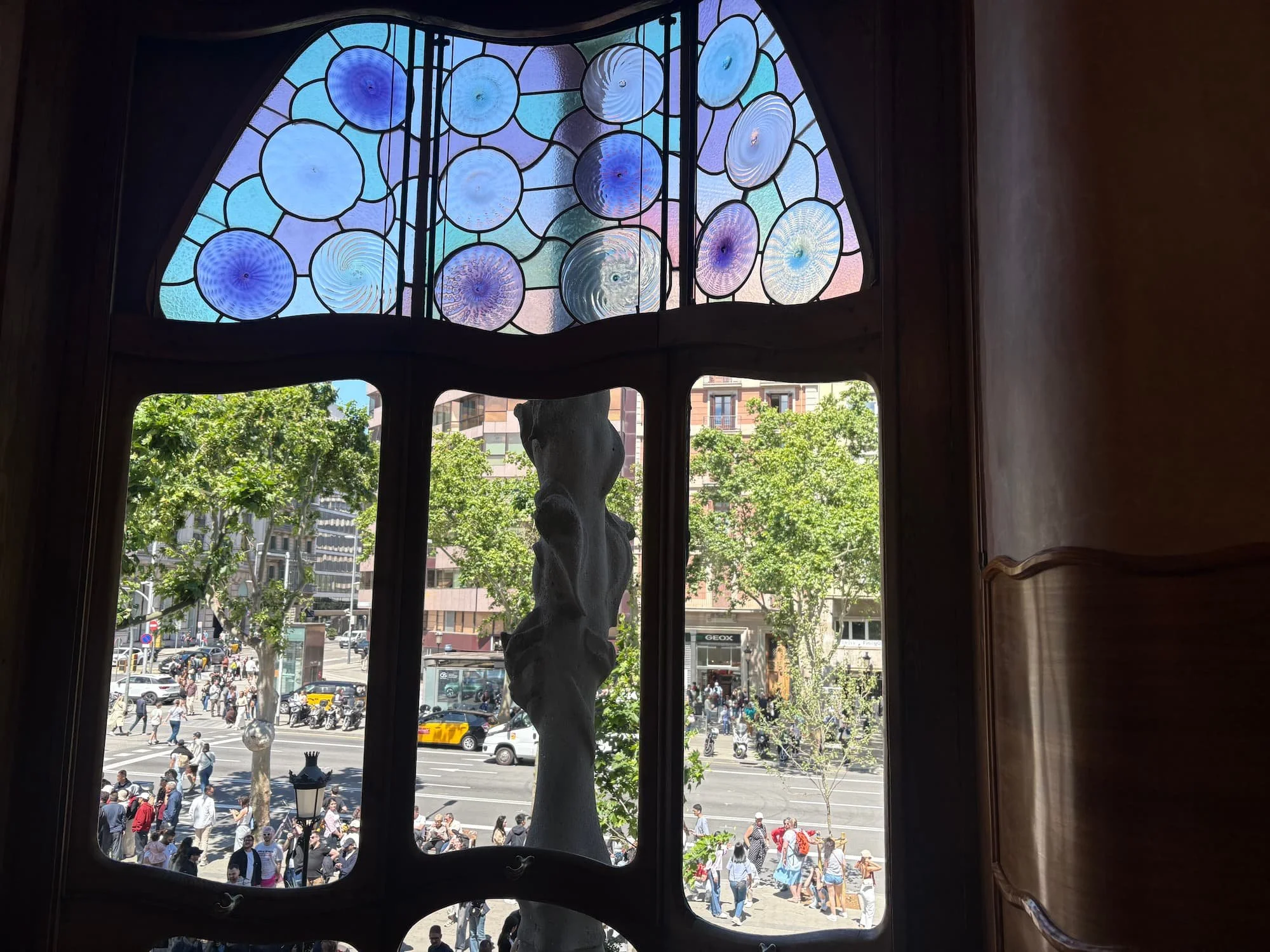 View through Casa Batlló's stained-glass bay window overlooking Passeig de Gràcia, featuring Gaudí's colourful circular glass designs and curved wooden frames.