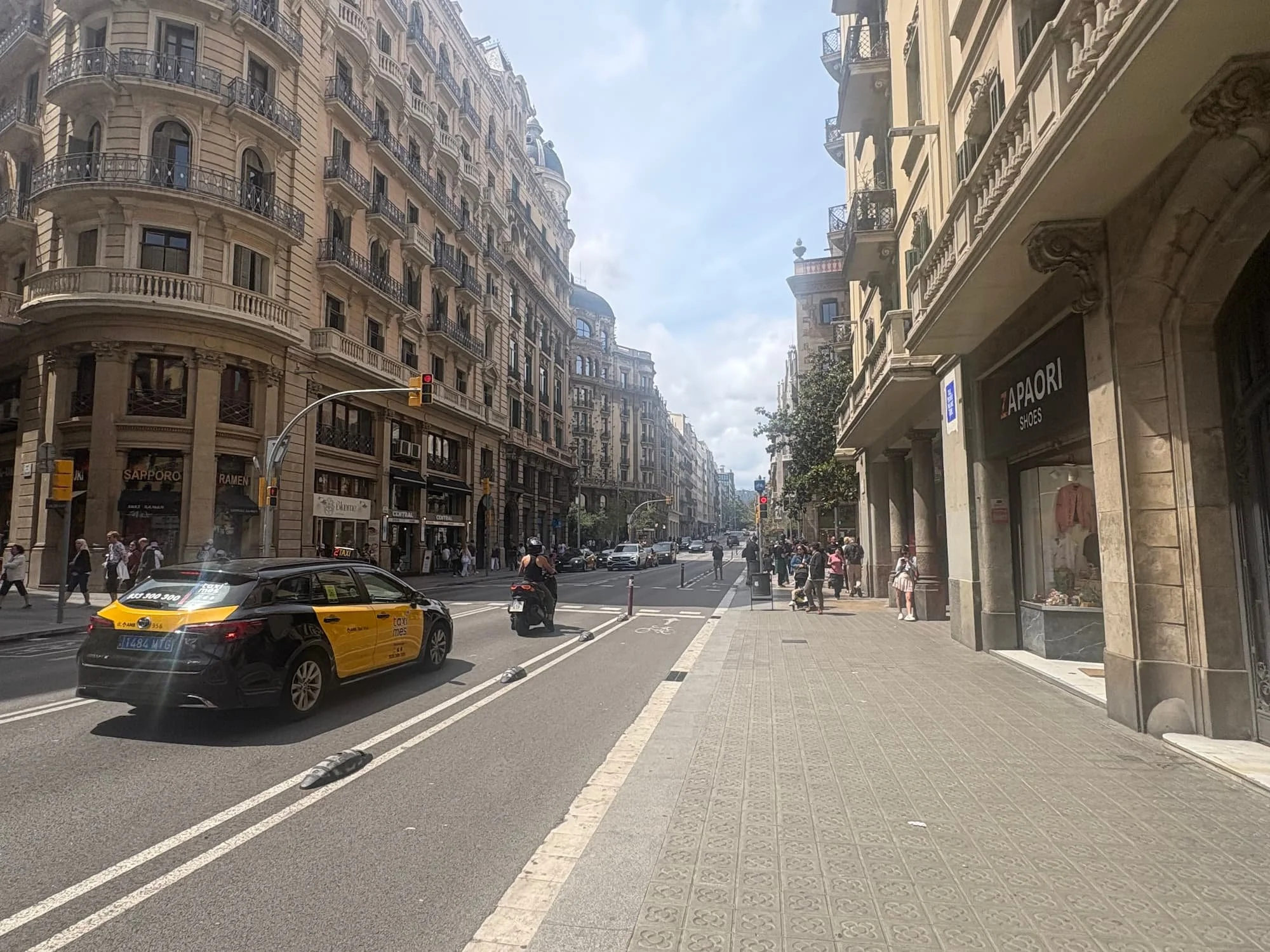 Wide boulevard in central Barcelona lined with historic apartment buildings, shops and cafés, showcasing the city's distinctive architecture and lively street atmosphere.