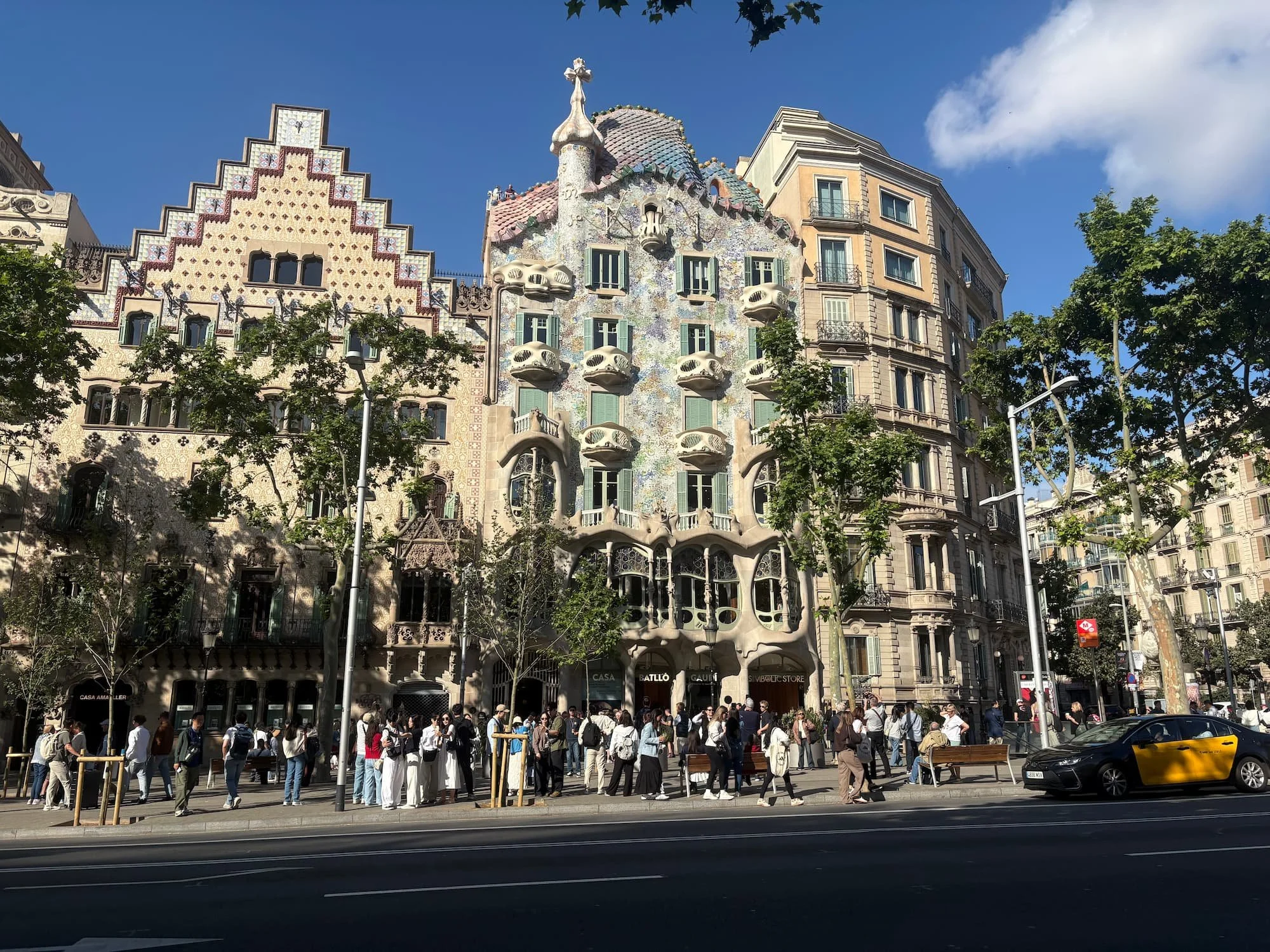 Wide street view of Casa Batlló on Passeig de Gràcia in Barcelona, showing Gaudí's colourful façade, dragon-inspired roof and crowds gathered outside.