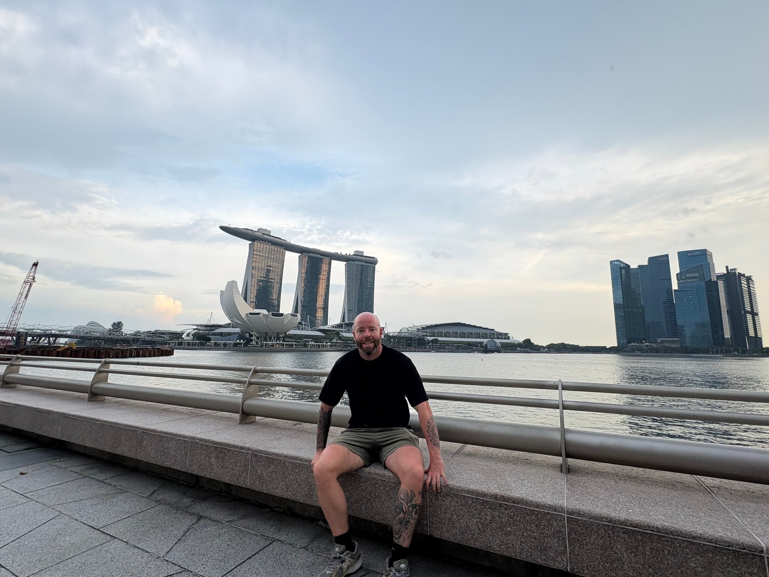 Tom Henty sitting along the Marina Bay waterfront with views of Marina Bay Sands and Singapore’s skyline in the background.