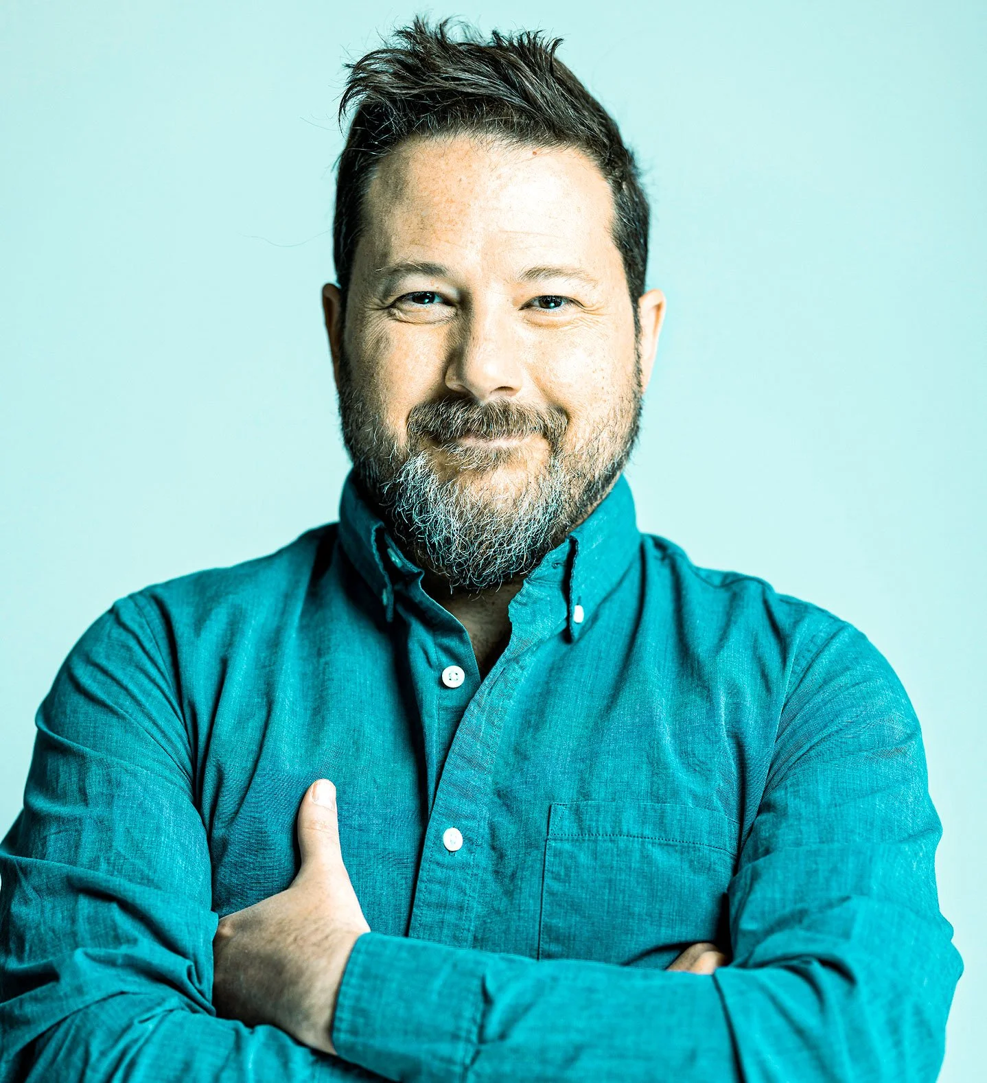 A man with short dark hair, a beard, and blue eyes smiling with arms crossed, wearing a teal button-up shirt against a light background.