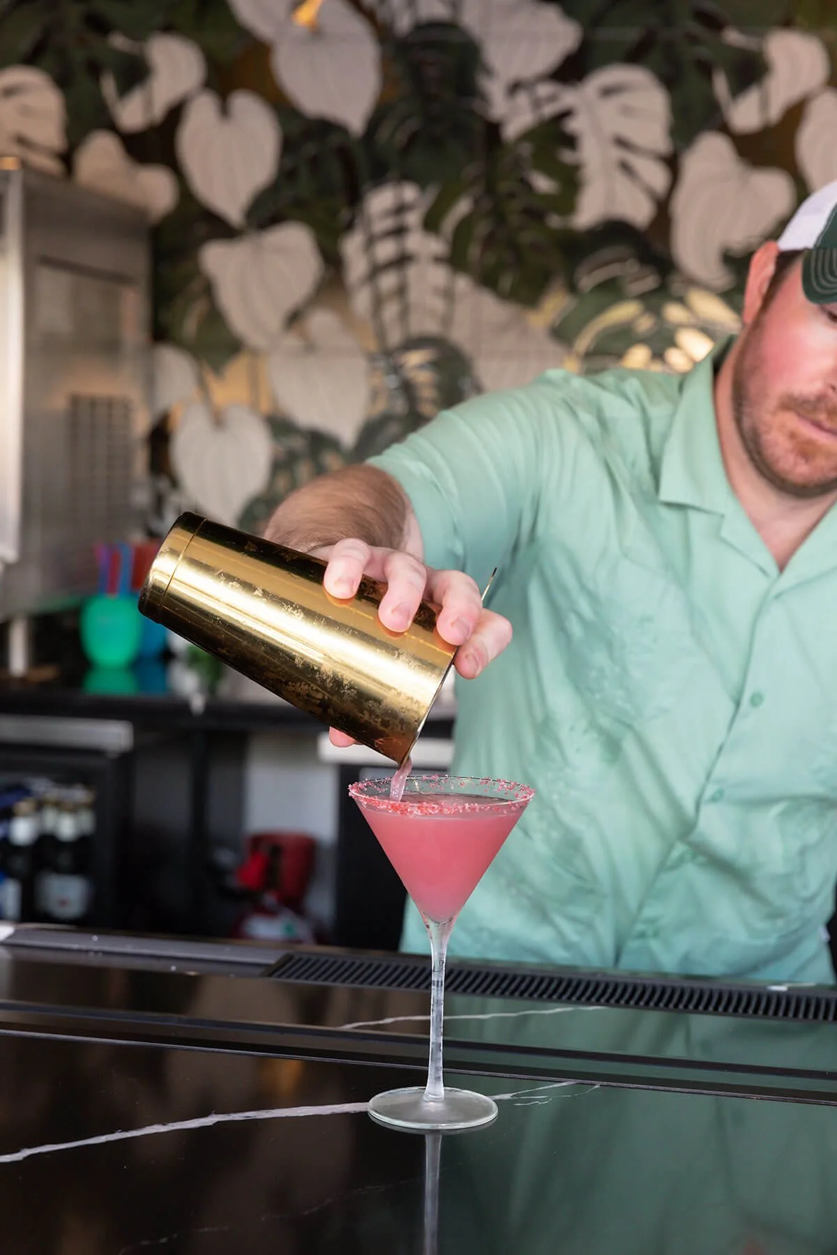 Bartender pouring pink cocktail into stemmed glass with sugared rim at bar with leafy wallpaper background.