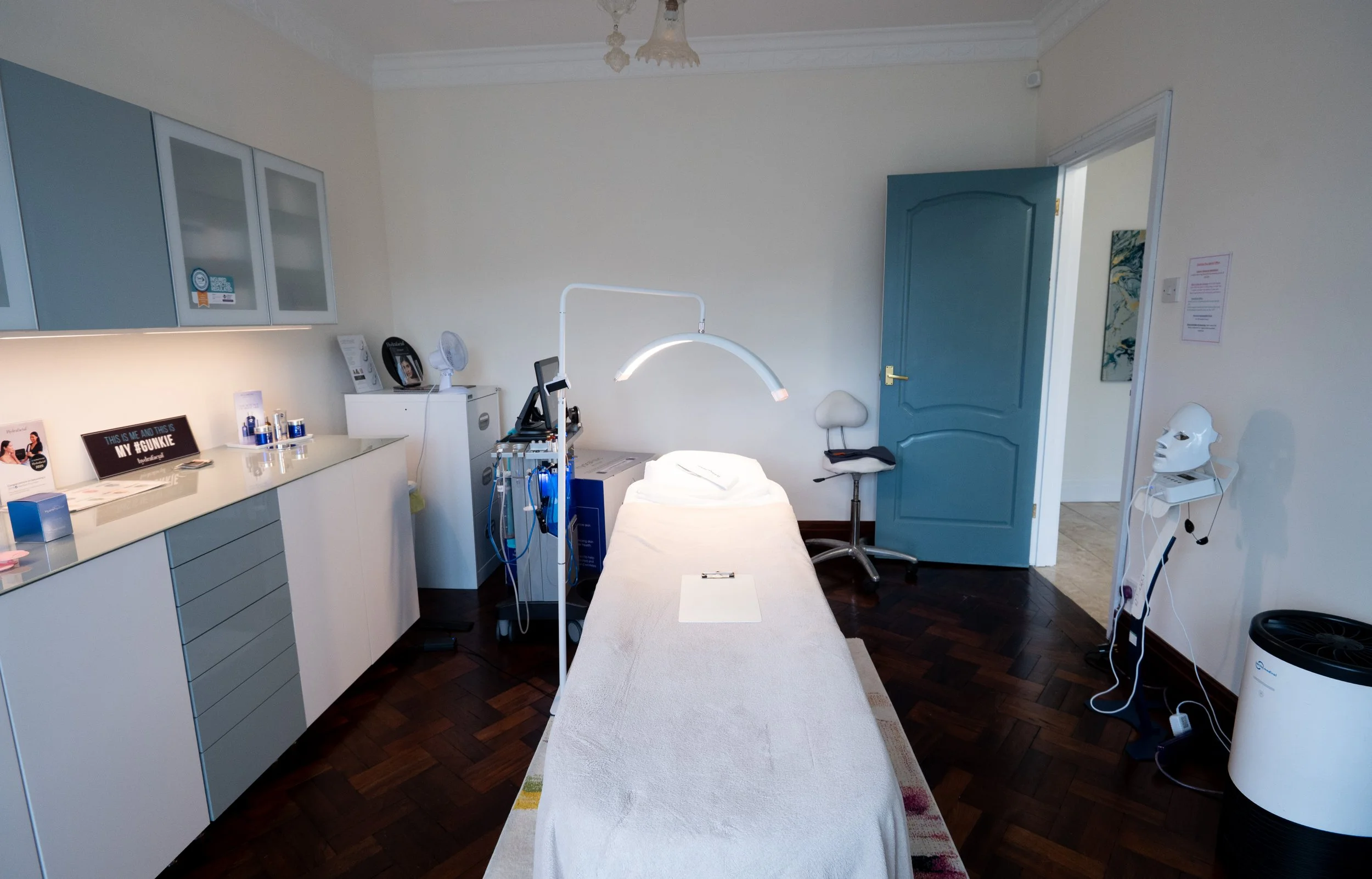 A medical examination room with a treatment bed in the center, medical equipment on the side, blue cabinets on the wall, a door open to another room, and various medical supplies and devices.