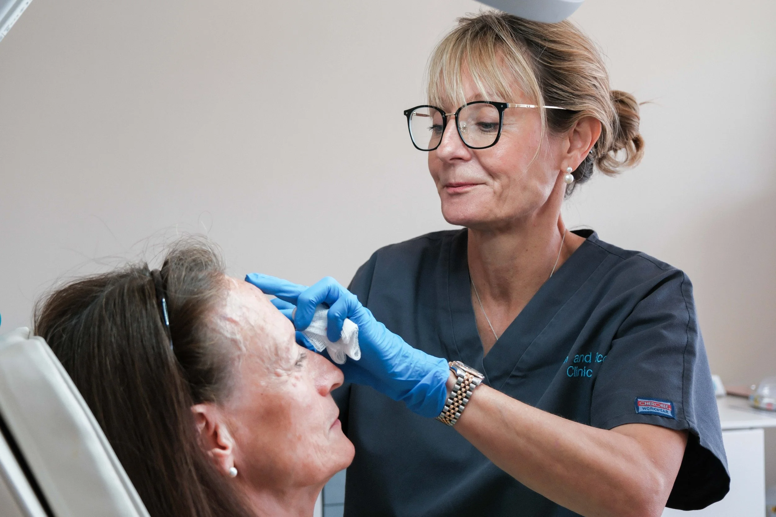 A healthcare professional wearing glasses, gloves, and a gray scrubs shirt examining an elderly woman's face in a clinical setting.