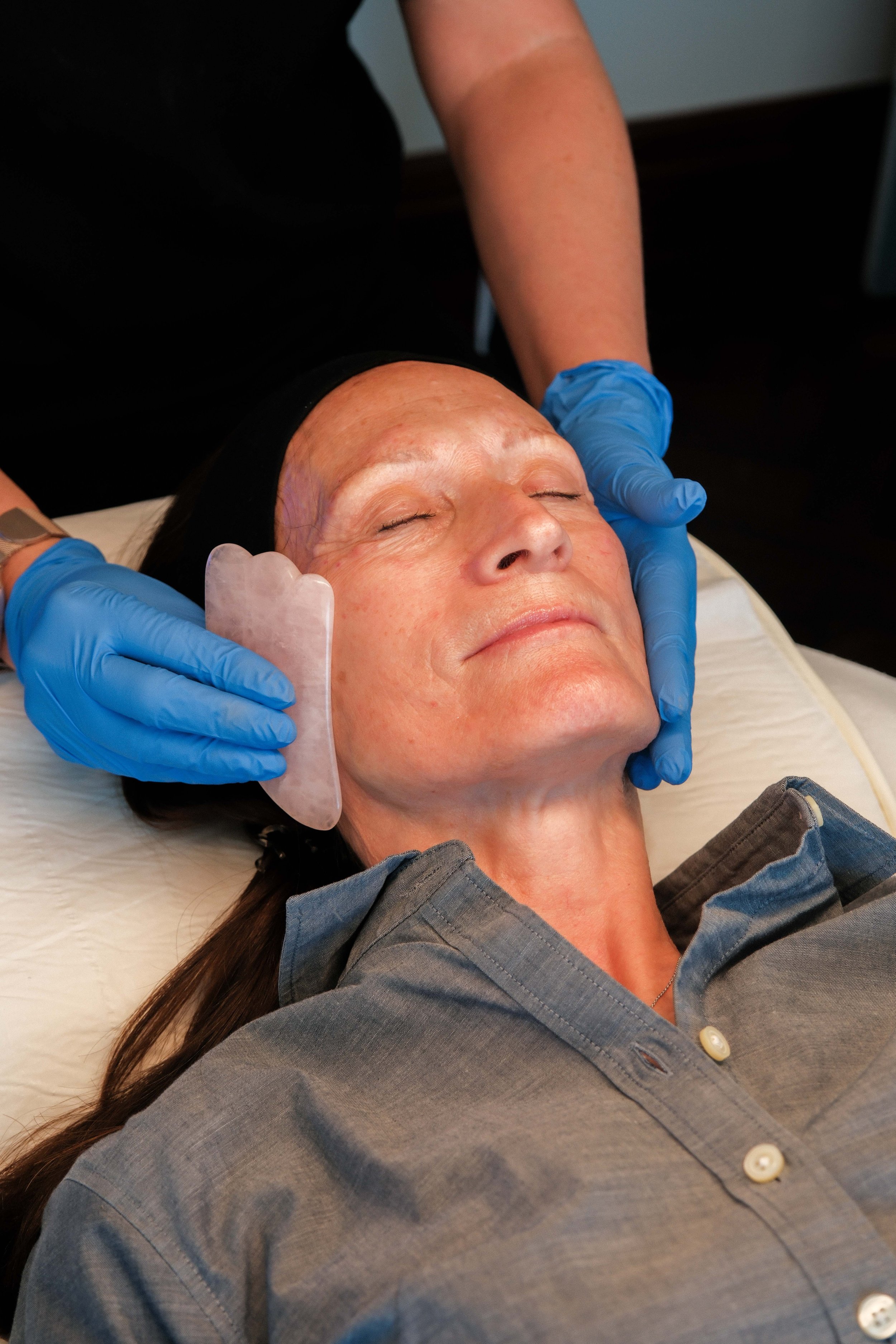 A woman receiving a facial treatment, lying on her back with eyes closed, while a skincare professional with blue gloves applies a facial mask.