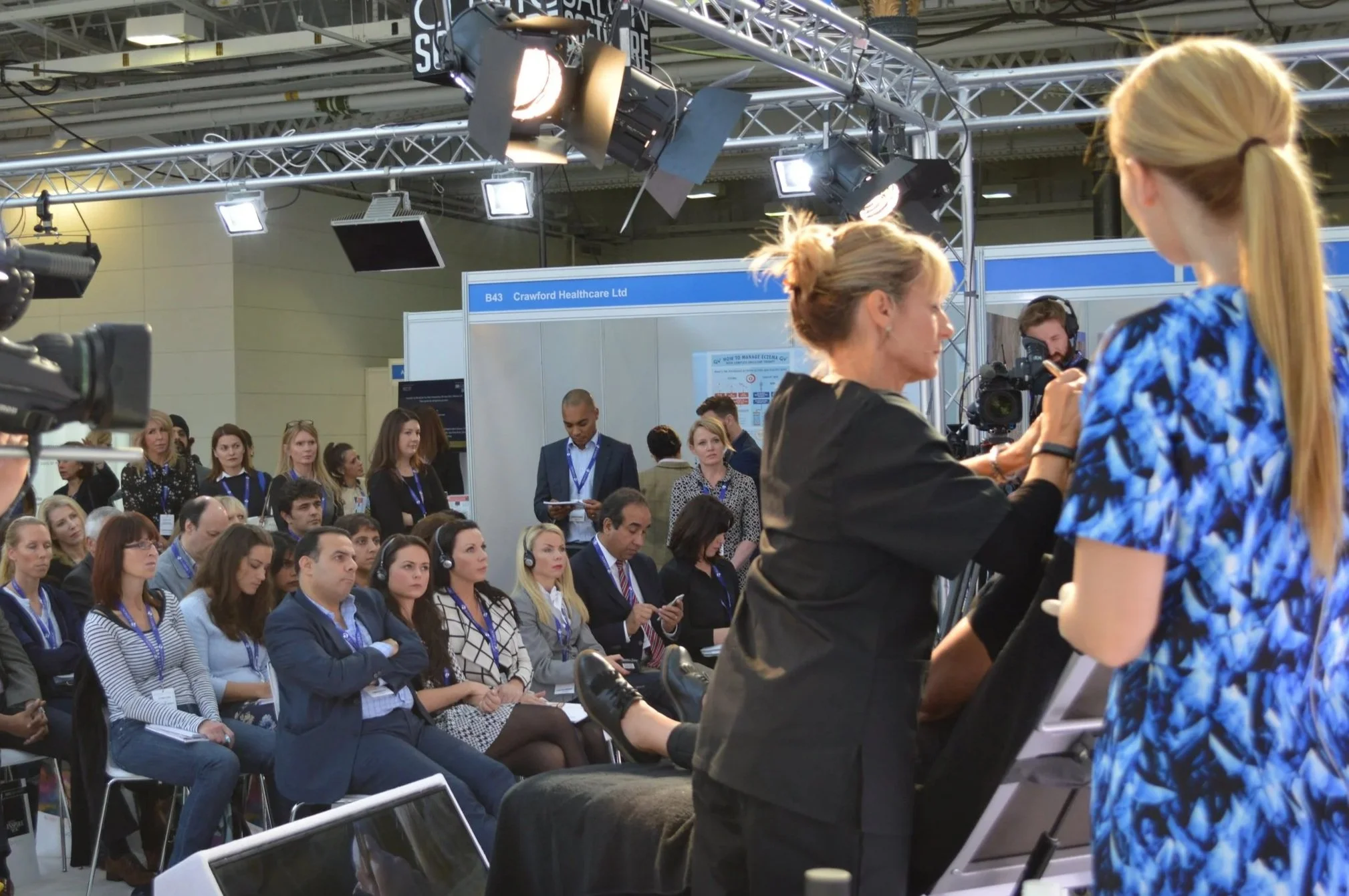 A group of journalists filming and interviewing a woman in a black coat at a conference or seminar. Audience members are seated and listening attentively, some taking notes or using headphones. The setting appears to be an indoor event space with technical equipment and signage.