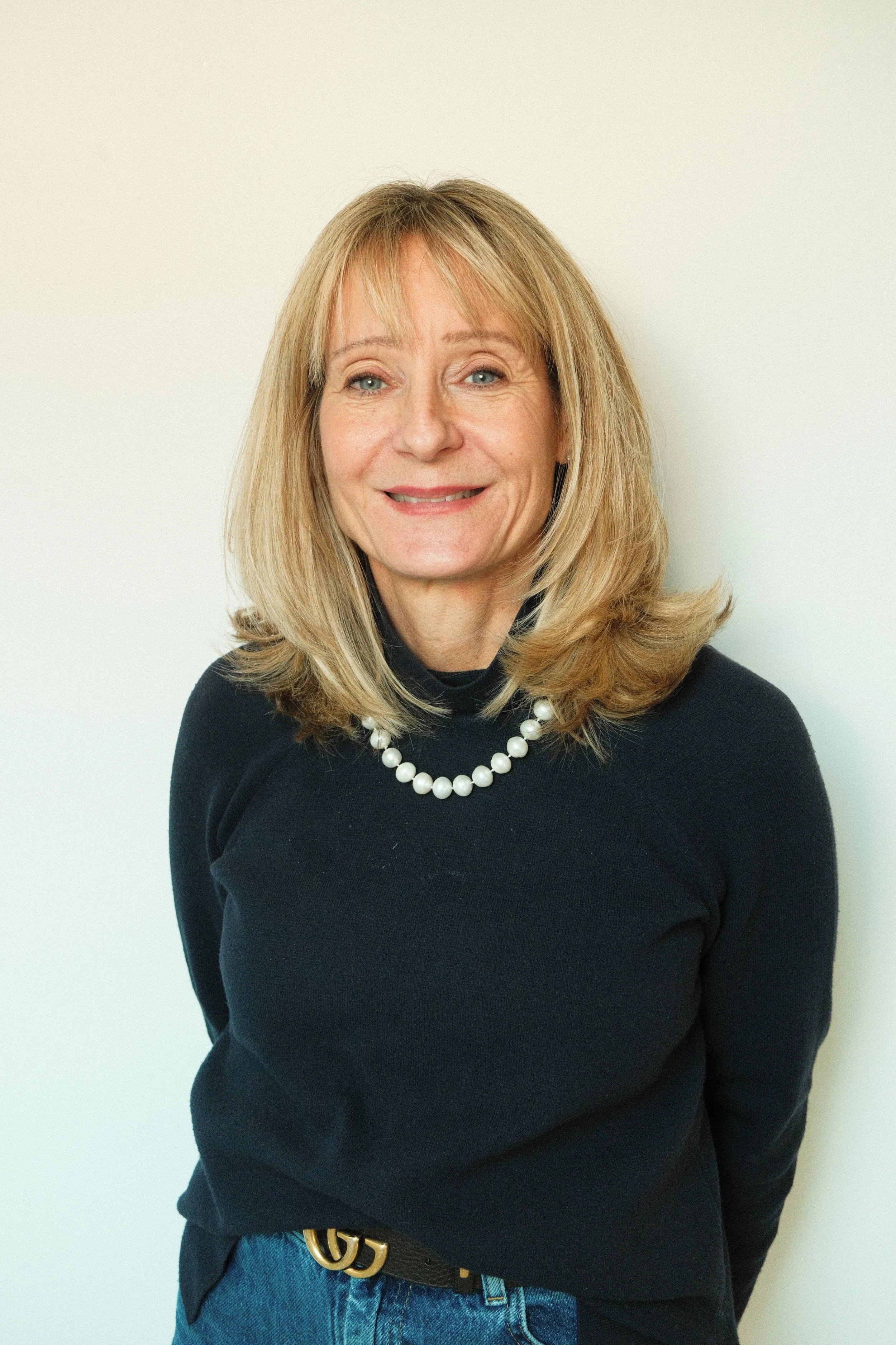 A middle-aged woman with blonde hair, wearing a black turtleneck and a pearl necklace, smiling and standing against a neutral background.