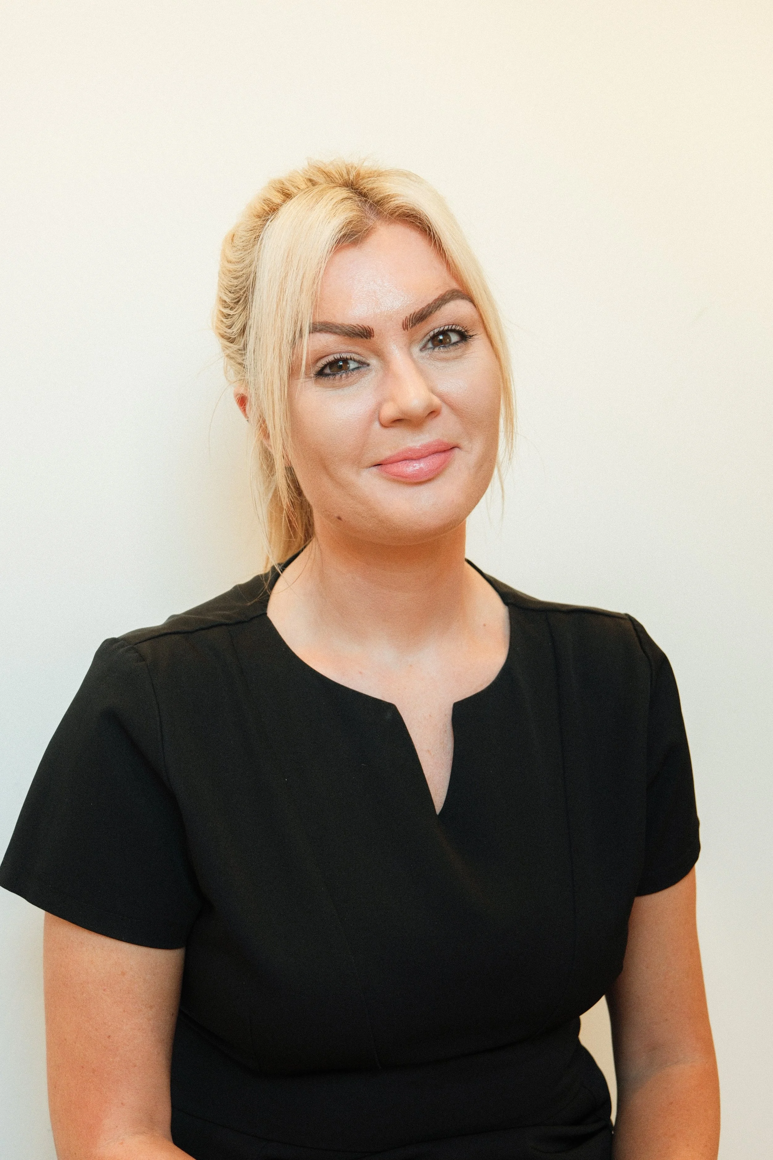 A woman with blonde hair pulled back, wearing a black top, smiling at the camera against a plain white background.