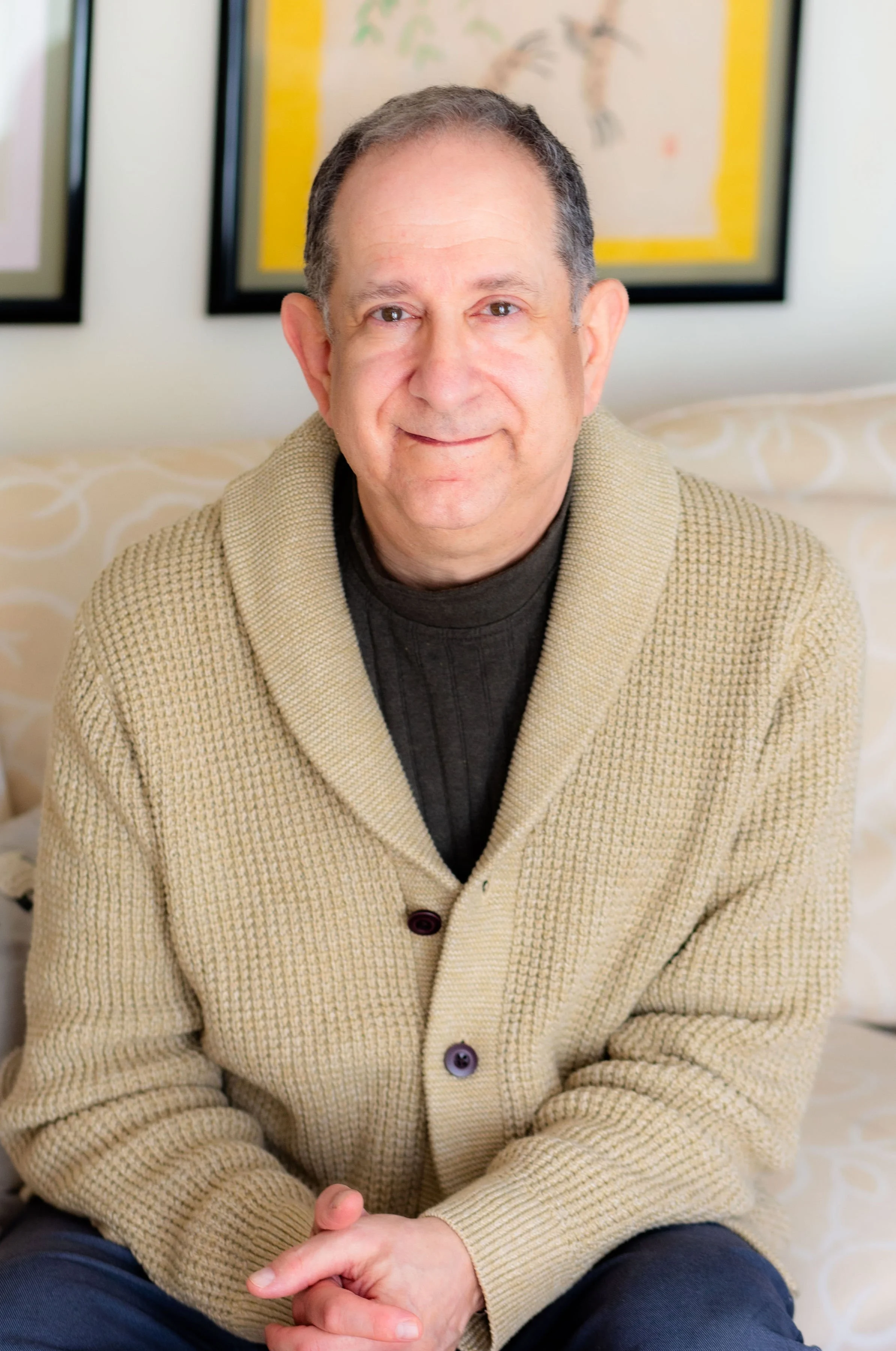 A middle-aged man with short, gray hair and light skin, smiling gently, sitting on a bed with patterned bedding. He is wearing a beige knit cardigan over a dark shirt, with hands loosely clasped in his lap. Behind him are framed pictures on the wall, possibly artwork or photographs.