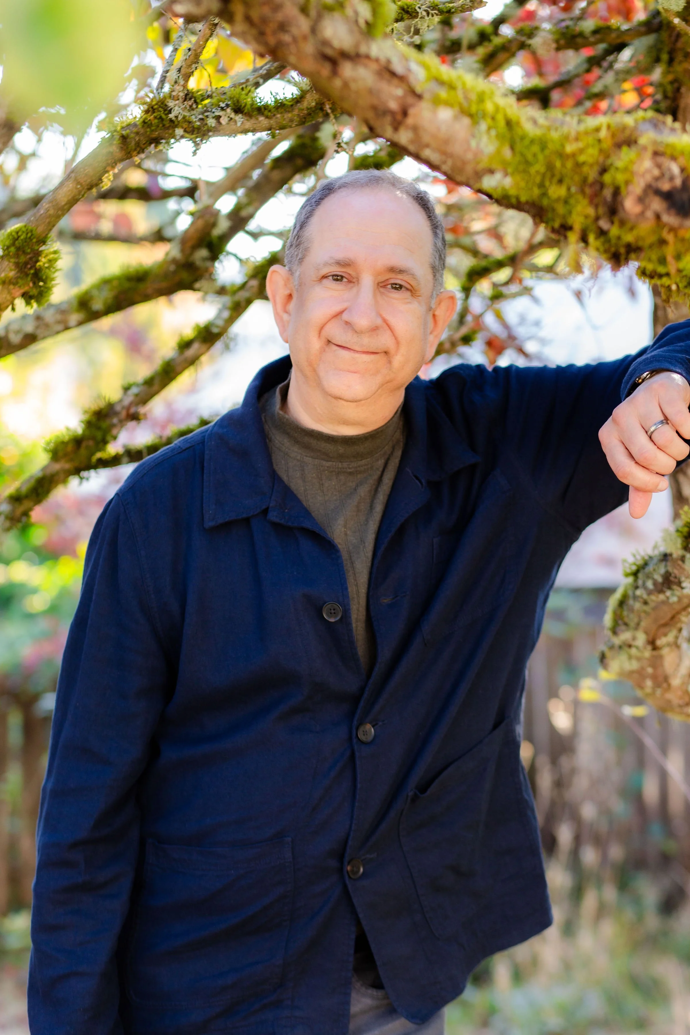 A middle-aged man with short gray hair smiling outdoors, standing among tree branches covered in moss and lichen, with blurred autumn leaves in the background.