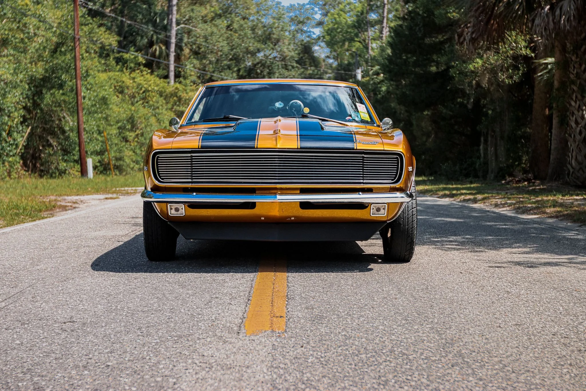 Front view of a vintage orange muscle car with black racing stripes parked on a rural road with trees on both sides.
