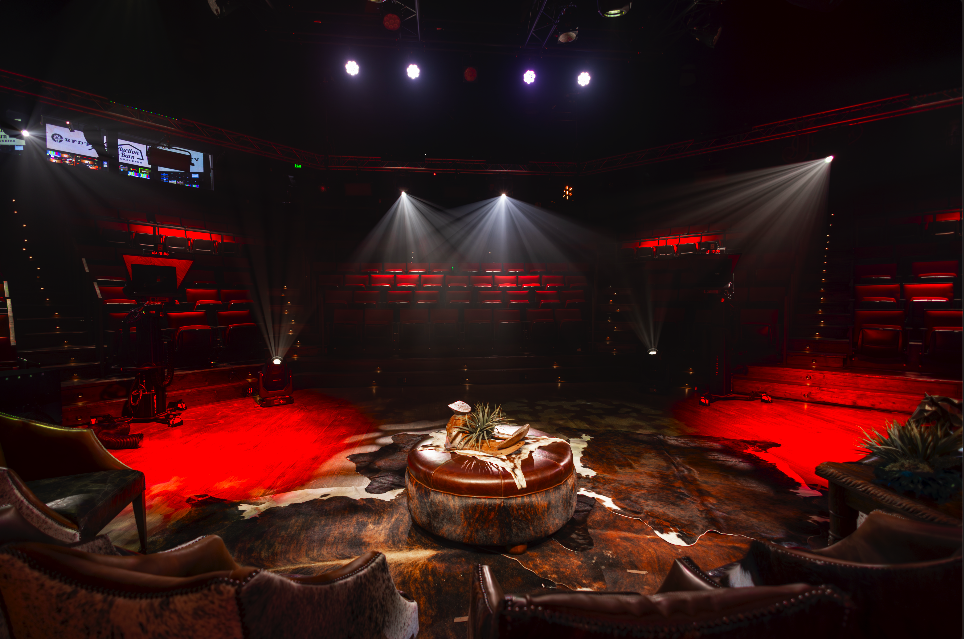 Empty theater or performance space with red seating, stage lighting, and a cowhide rug with a round ottoman in the center, decorated with plants.