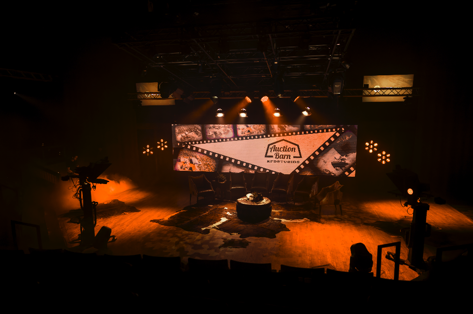 Stage set with chairs, a small round table, and a backdrop displaying a logo for Auction Barn.