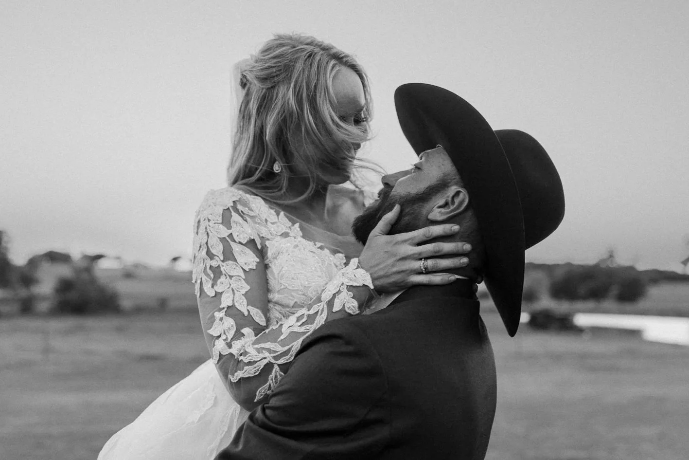 A black and white photo of a man with a beard wearing a wide-brimmed hat, holding a woman in a lace dress, as they gaze into each other's eyes outdoors.