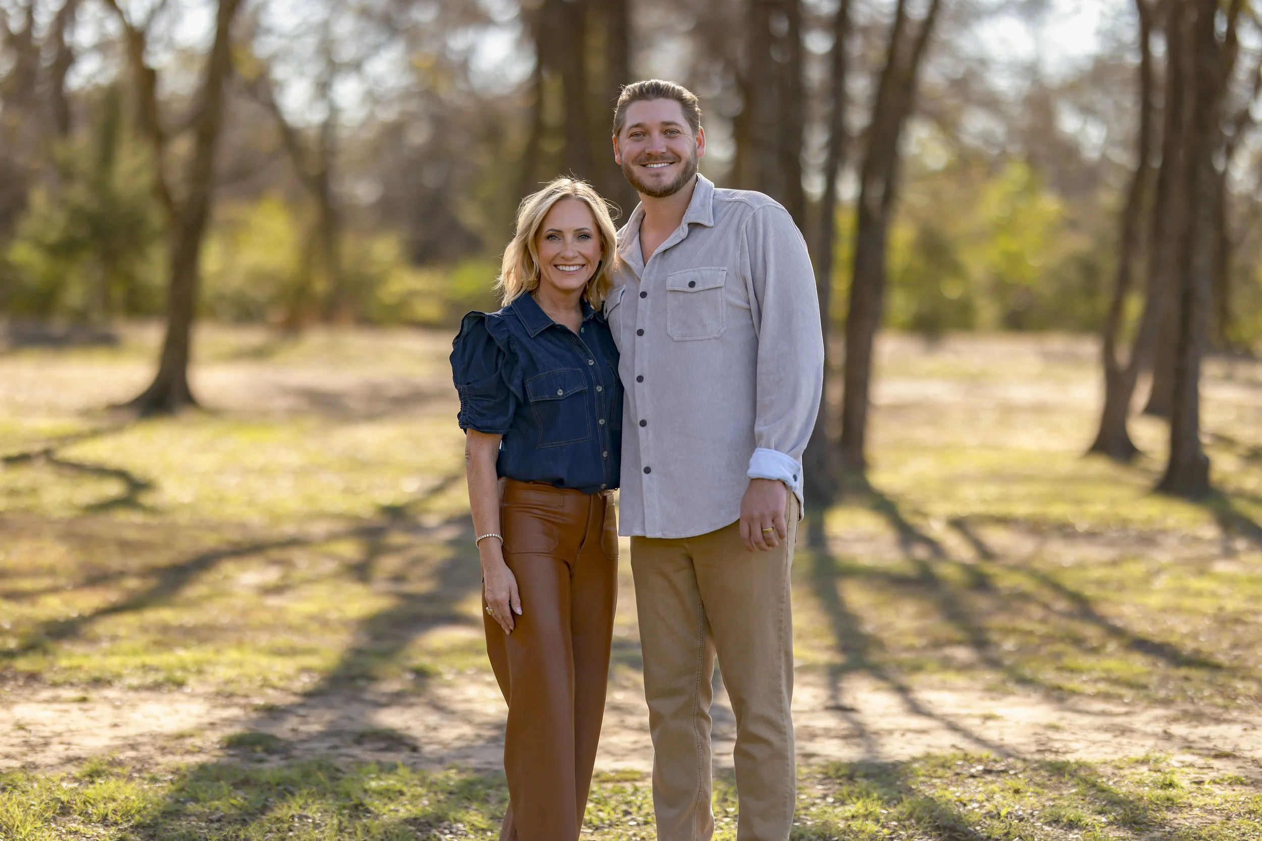 A happy couple standing outdoors in a park with trees in the background during autumn. The woman has blonde hair, is wearing a dark blue shirt and brown pants, and the man has short hair, a beard, is wearing a light gray button-down shirt and beige p